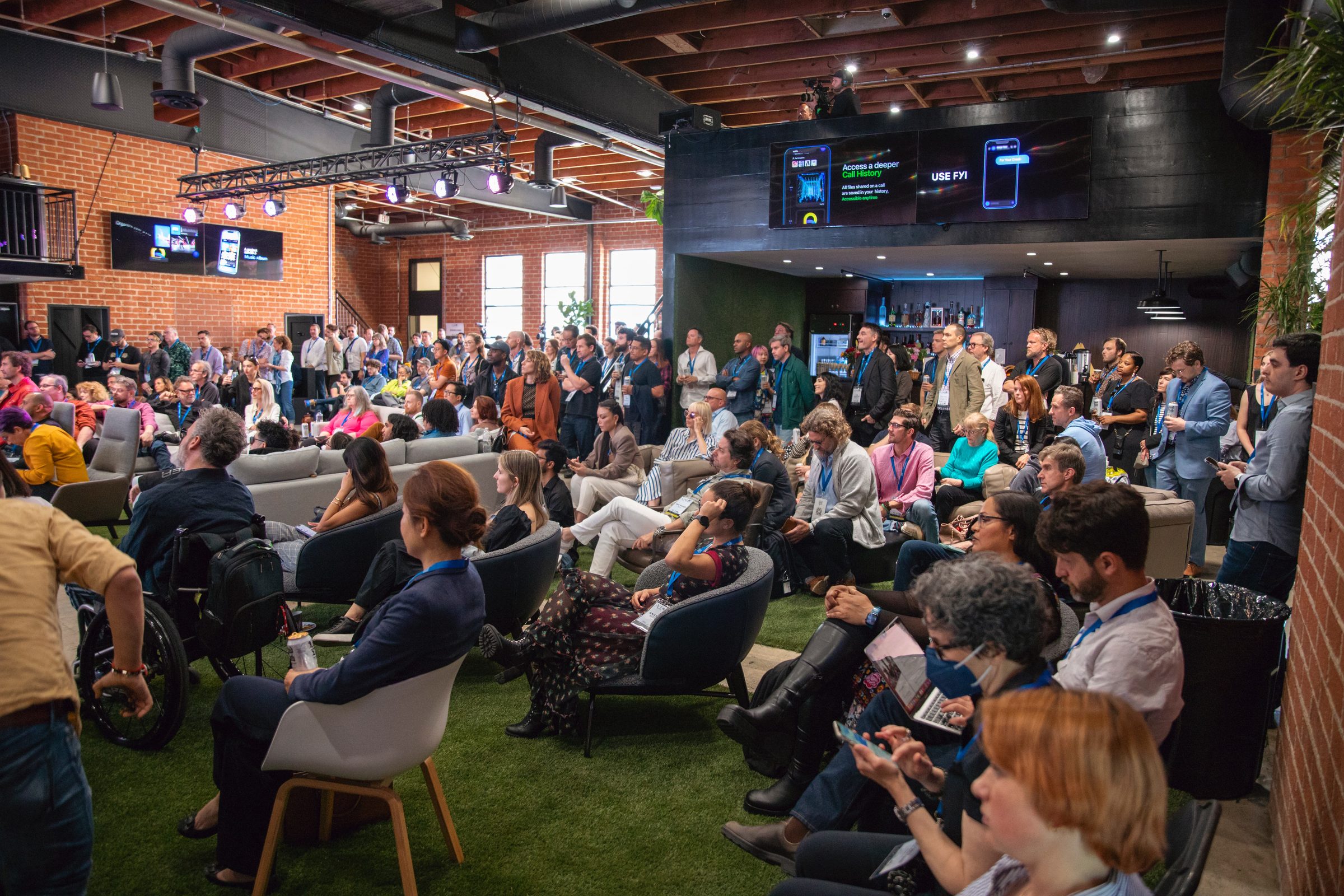 A large audience seated on the floor and couches in a brick studio with plants and a lighting rig.