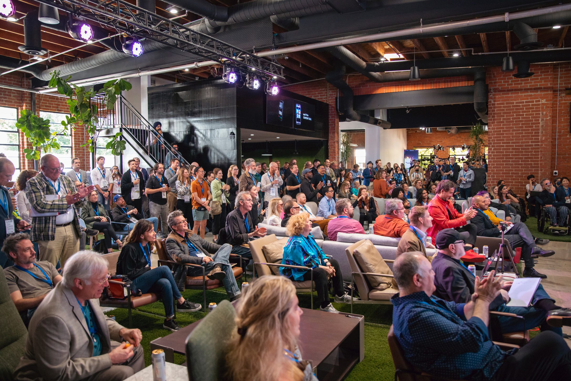 Attendees fill a brick-walled studio, seated close together and facing the stage.