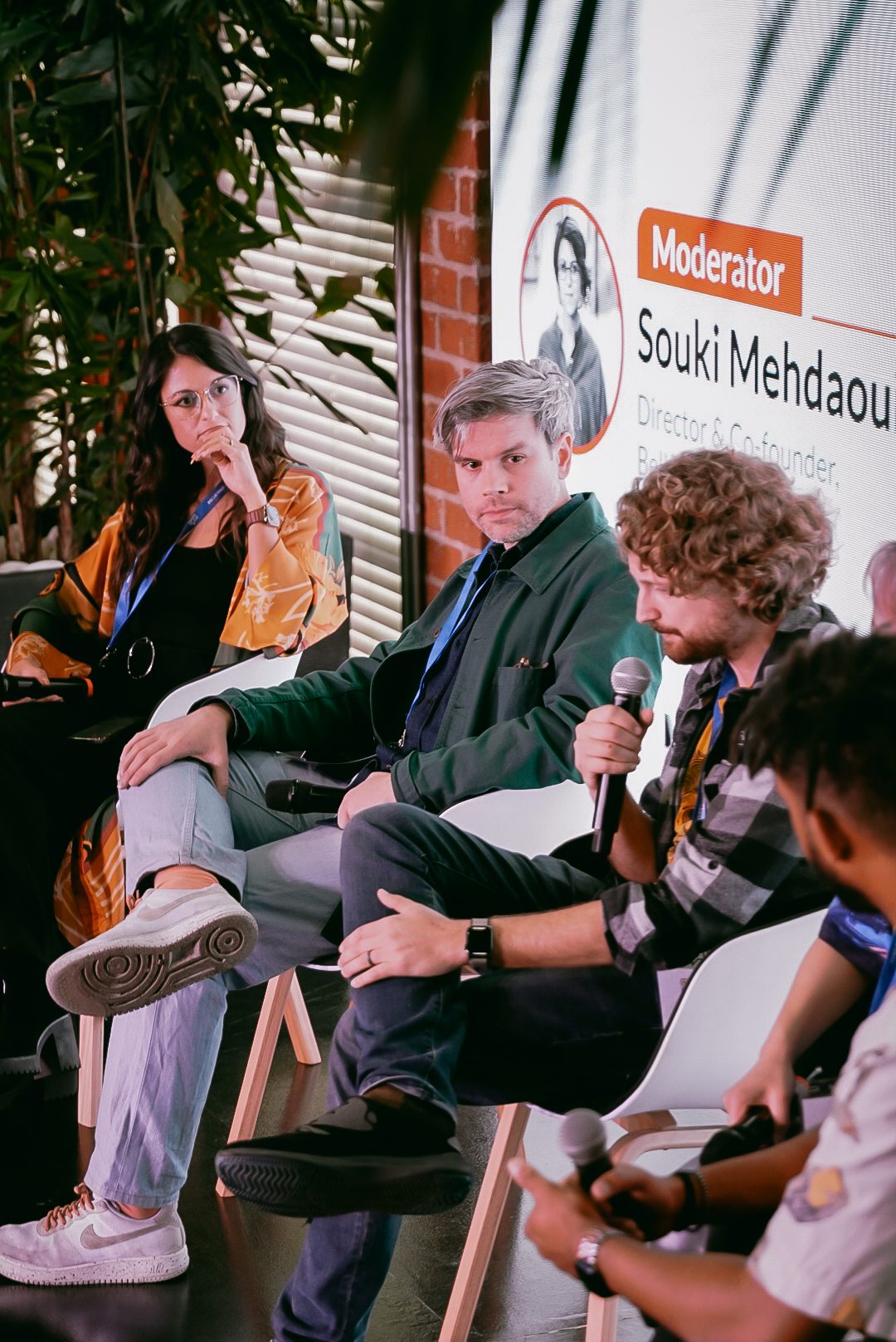 Three panelists seated on stage, Souki Mansoor named as moderator on the screen behind them.