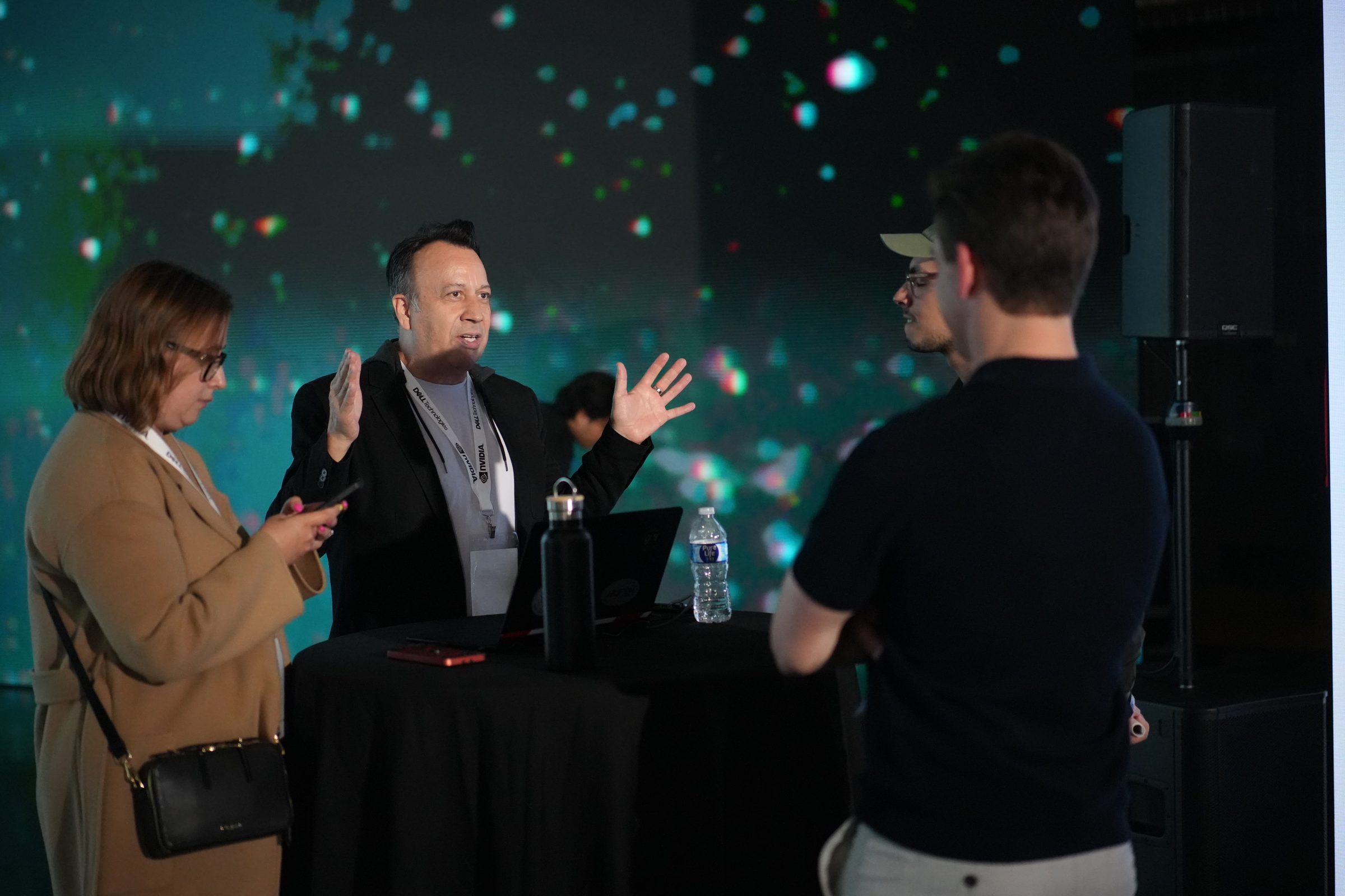 Three attendees talk in front of a dark stage wall filled with projected sparks of light.