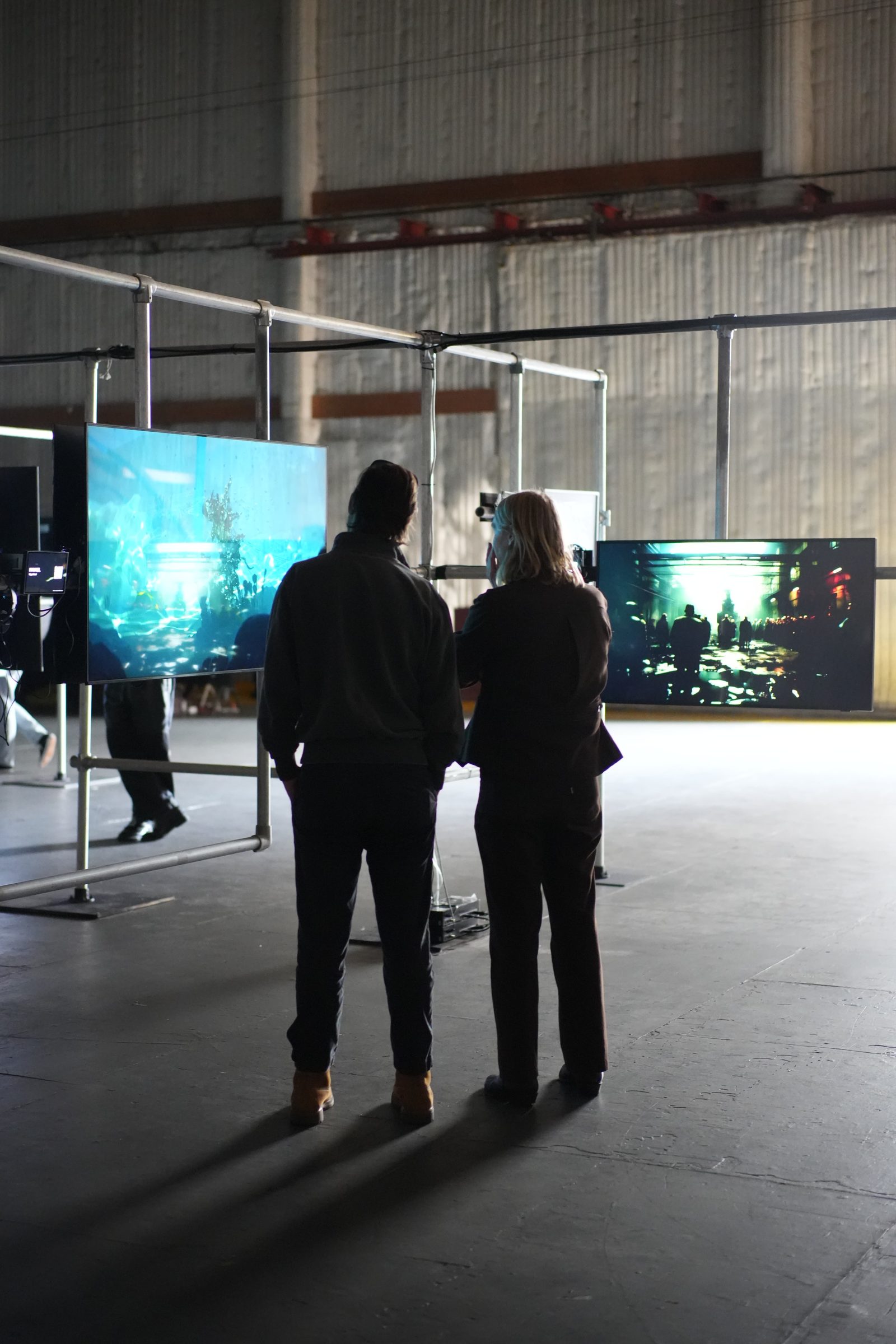 Two attendees stand in a dark gallery facing a row of screens of generated artwork.