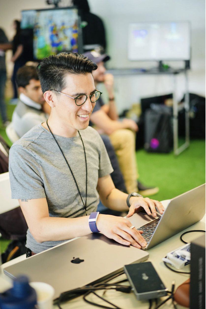 Minh Do smiles at a laptop in the Gen Lab, screens and a small crowd behind him.