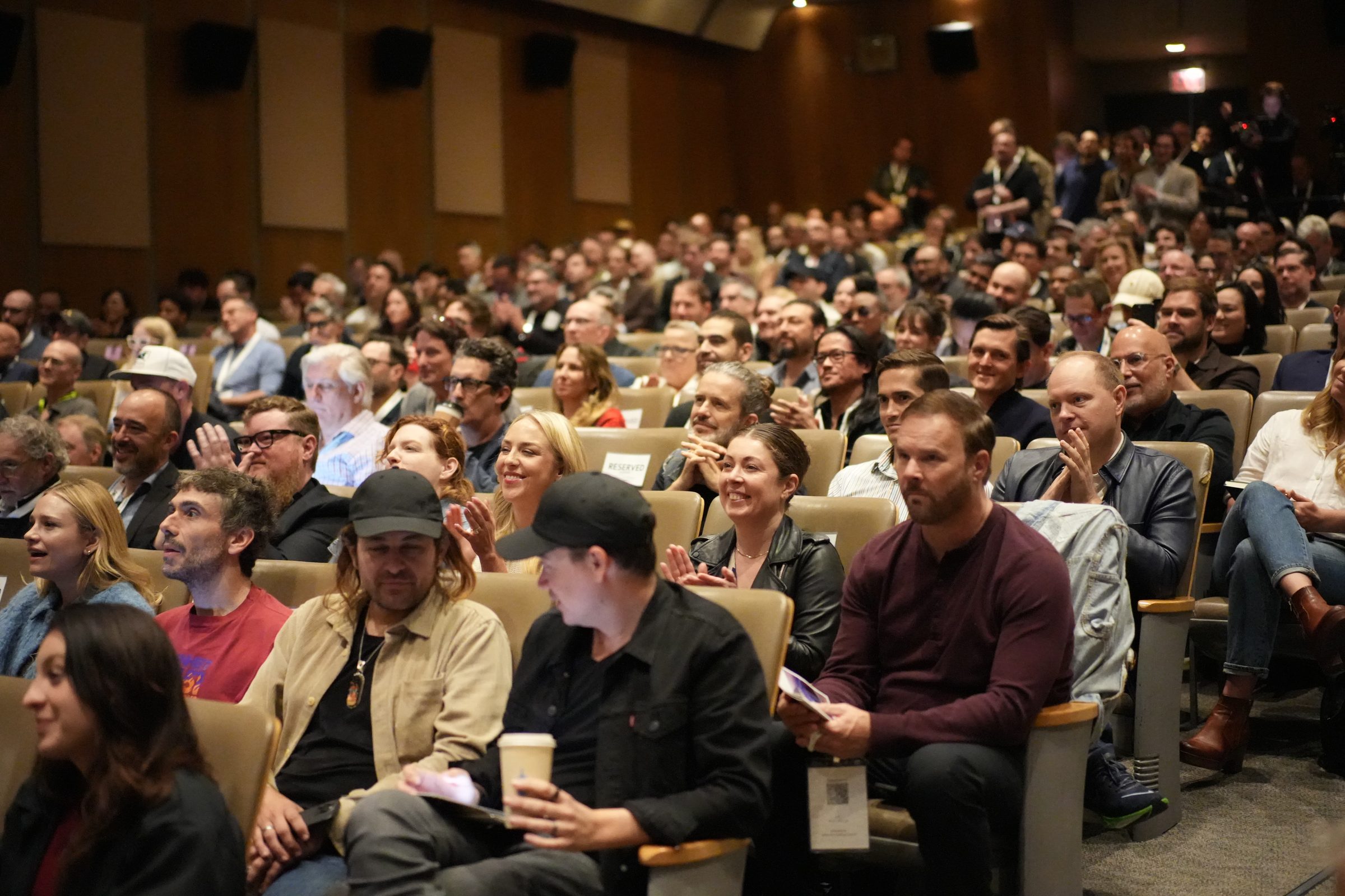 A full auditorium of attendees watches a panel, many leaning forward in their seats.