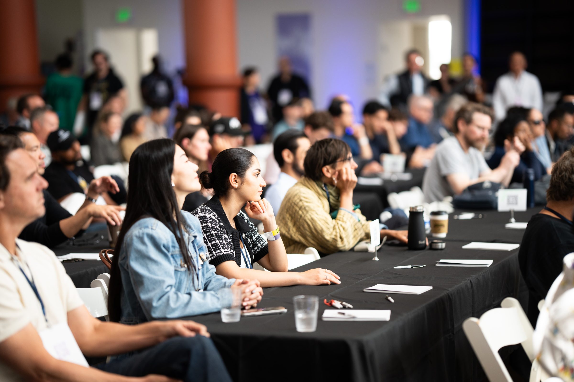 A packed workshop, attendees seated along a long black table, facing the front of the room.