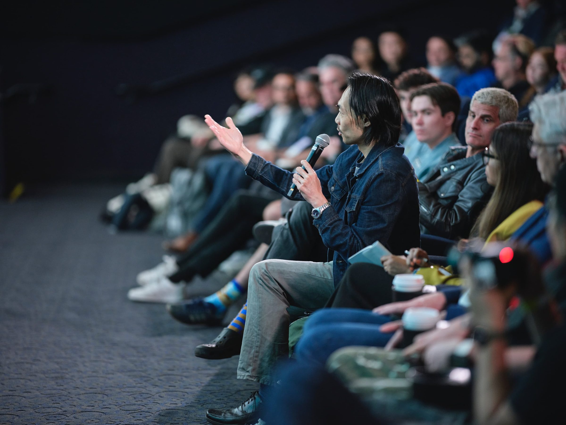 A seated attendee gestures with an open hand while asking a question into a microphone.