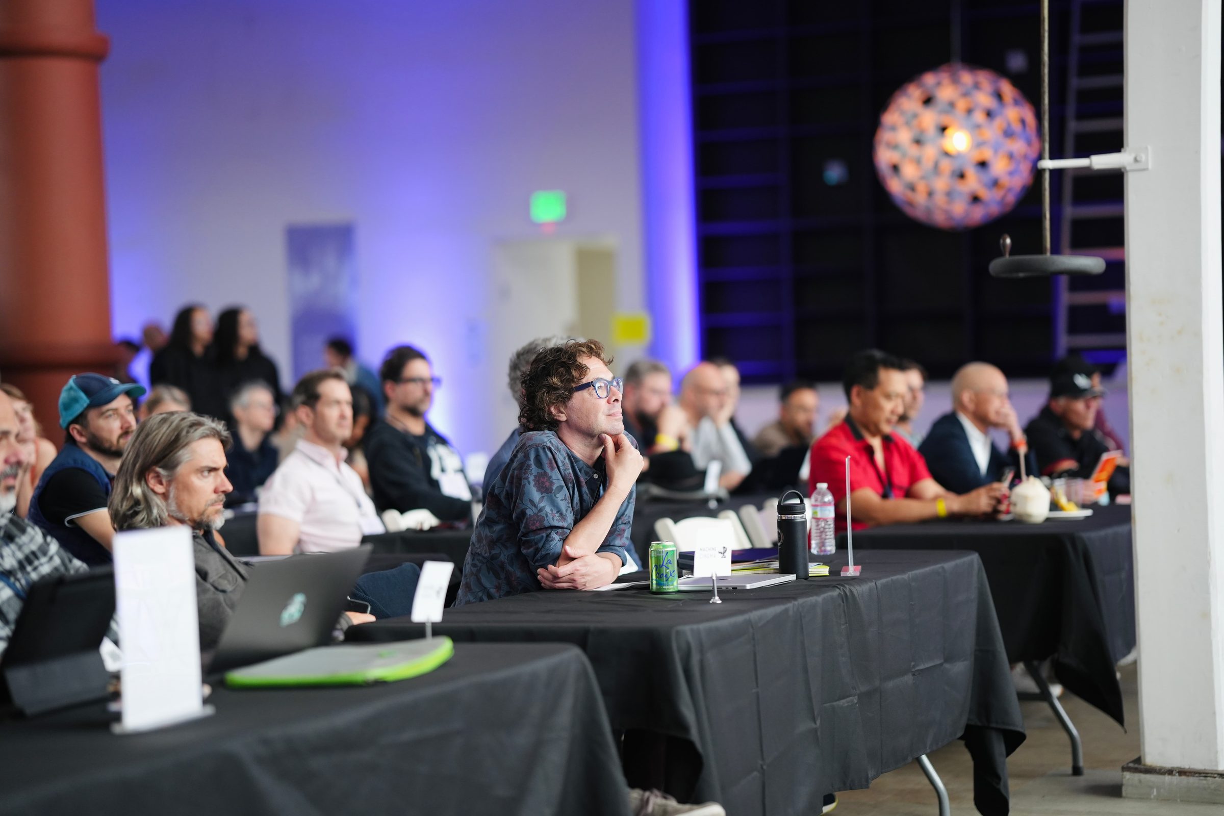 Rows of attendees at black-draped tables with laptops, under purple light and a hanging lamp.