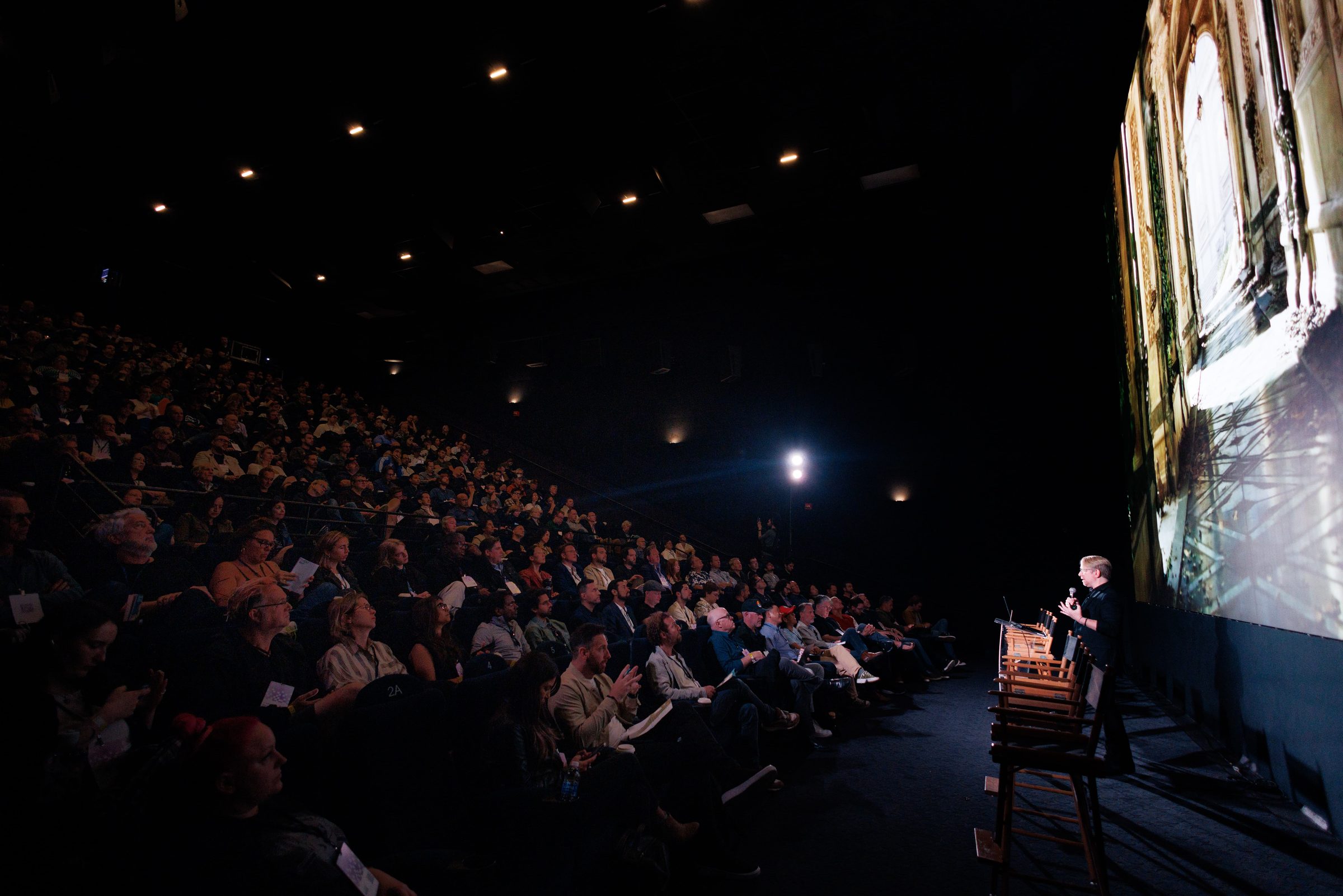 A full, dark Culver Theater watches Rick Champagne on stage, his presentation filling the screen.