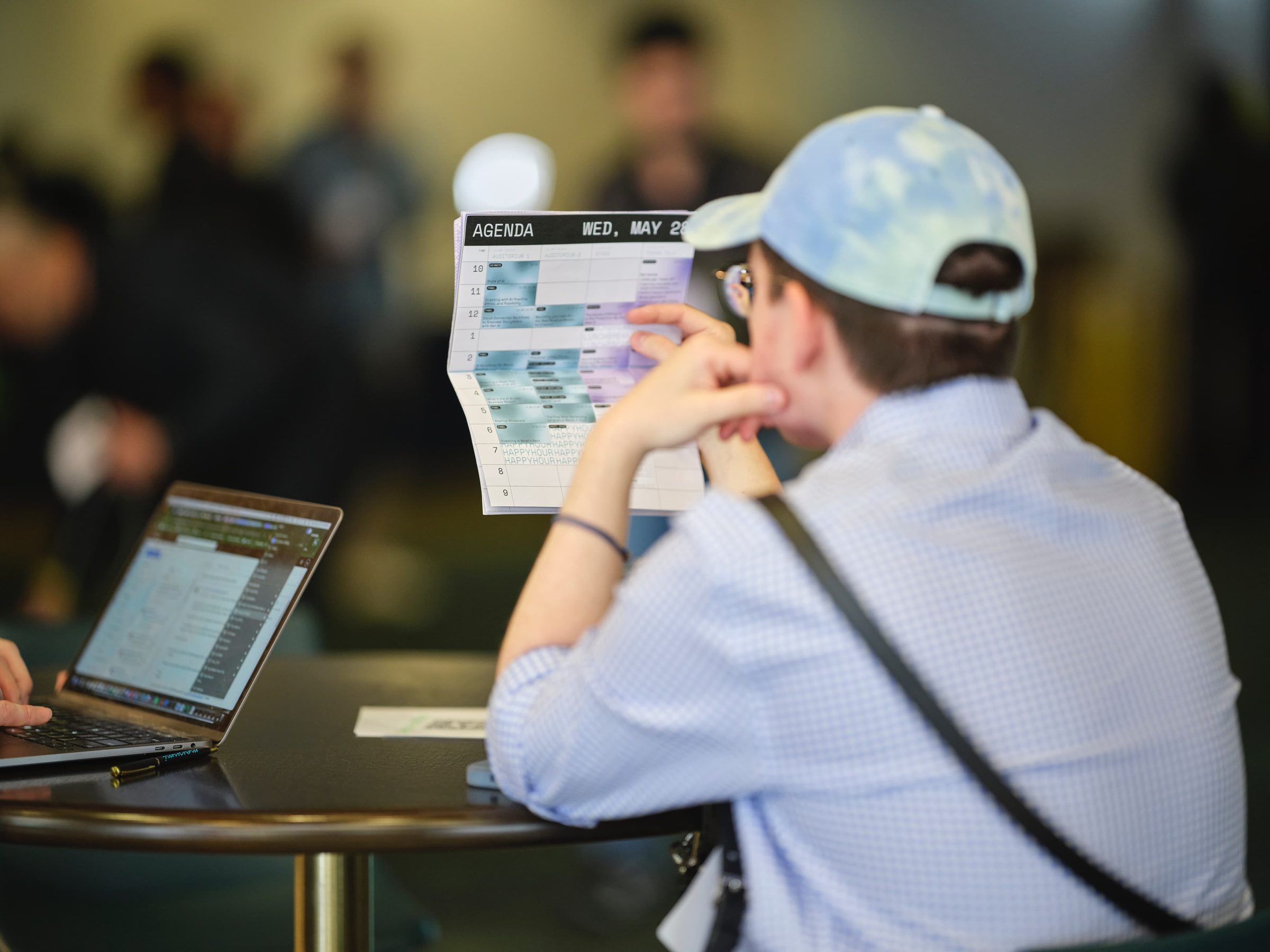 An attendee in a cap holds up a folded conference schedule beside an open laptop.