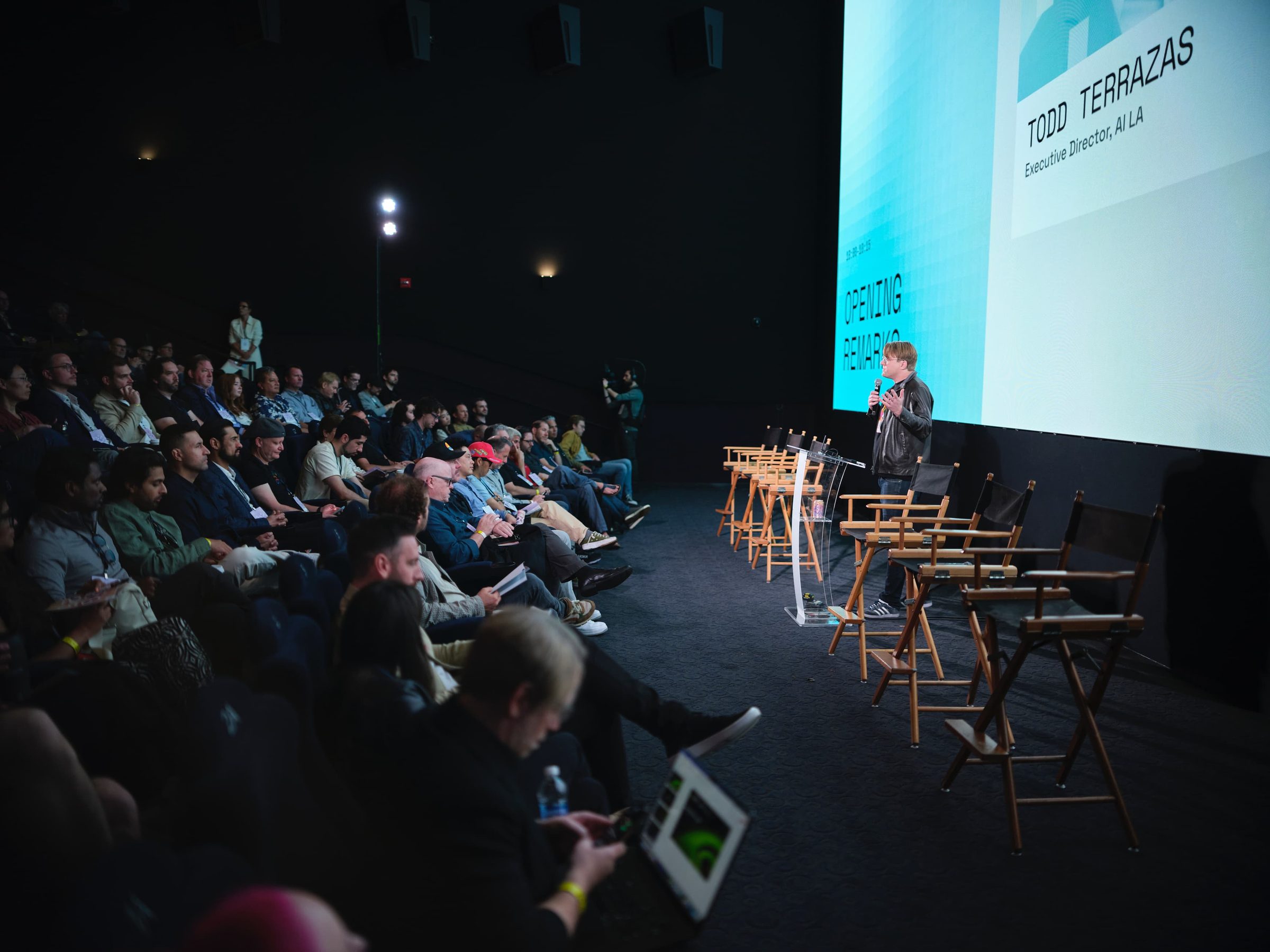 Todd Terrazas speaks from the stage of a dark theater, rows of director's chairs beside him.