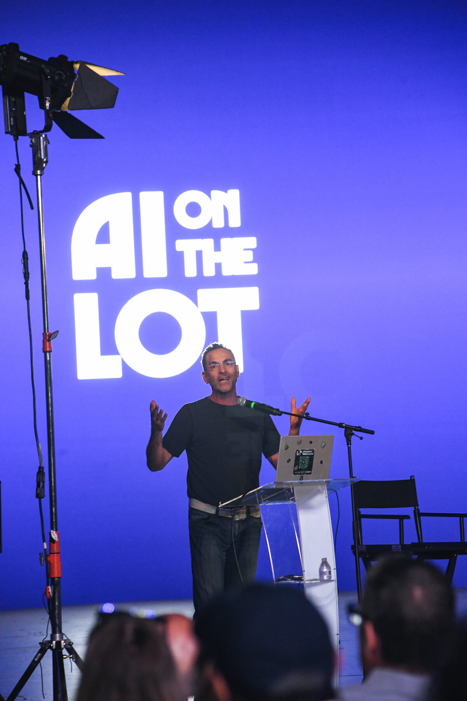 Adi Sideman throws his hands up mid-pitch at the lectern, the AI on the Lot logo glowing on the blue wall.