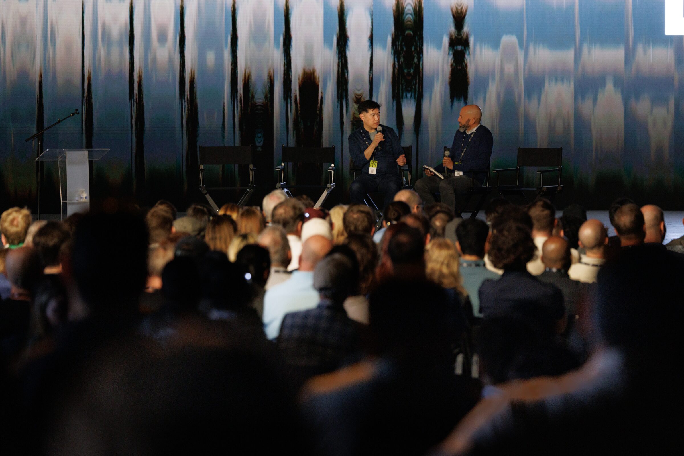 Albert Cheng talks with a fellow speaker in director's chairs on stage, seen past a packed, out-of-focus crowd.