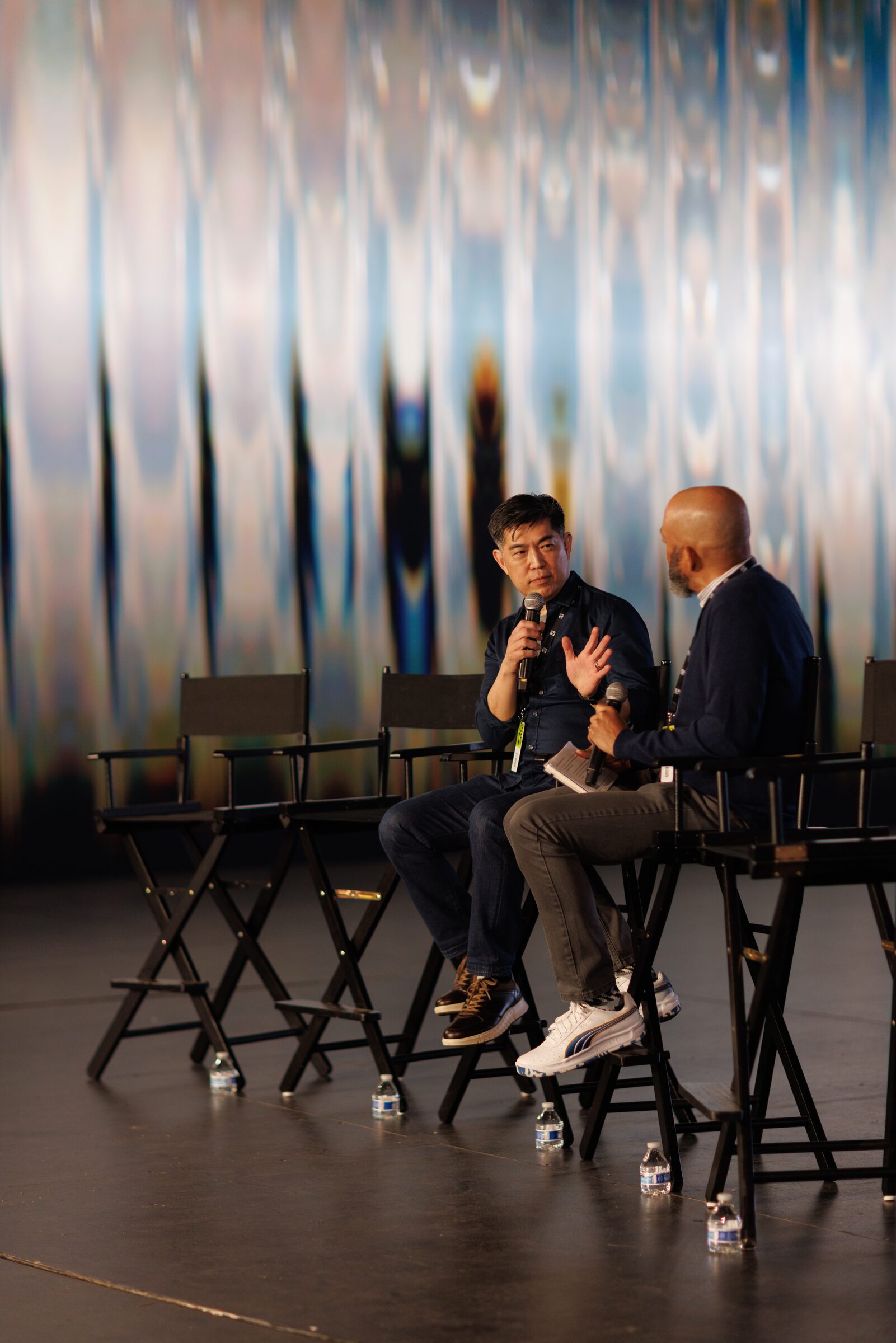 Albert Cheng speaks into a mic from a tall director's chair beside a second speaker, the projected wall above.