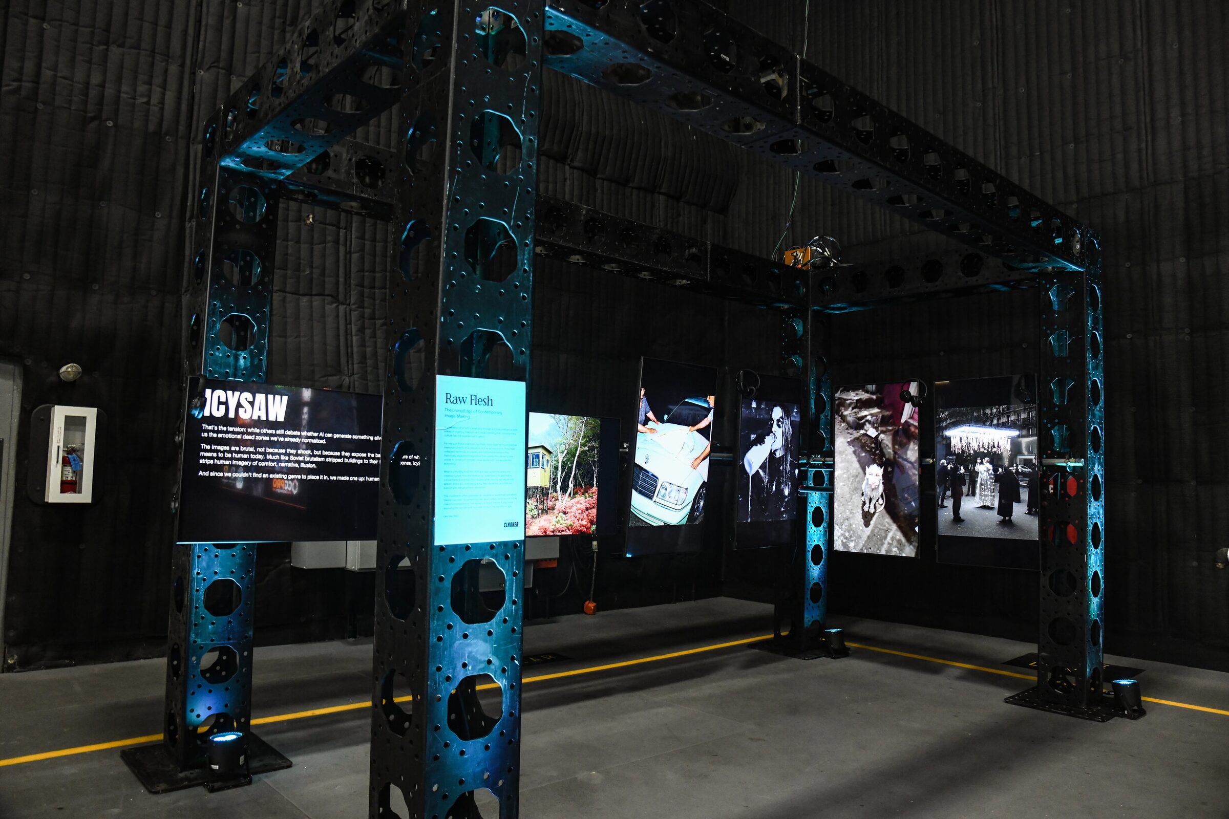 Clanker Magazine's art display: framed screens and placards hung on a blue-lit steel truss in a dark stage.