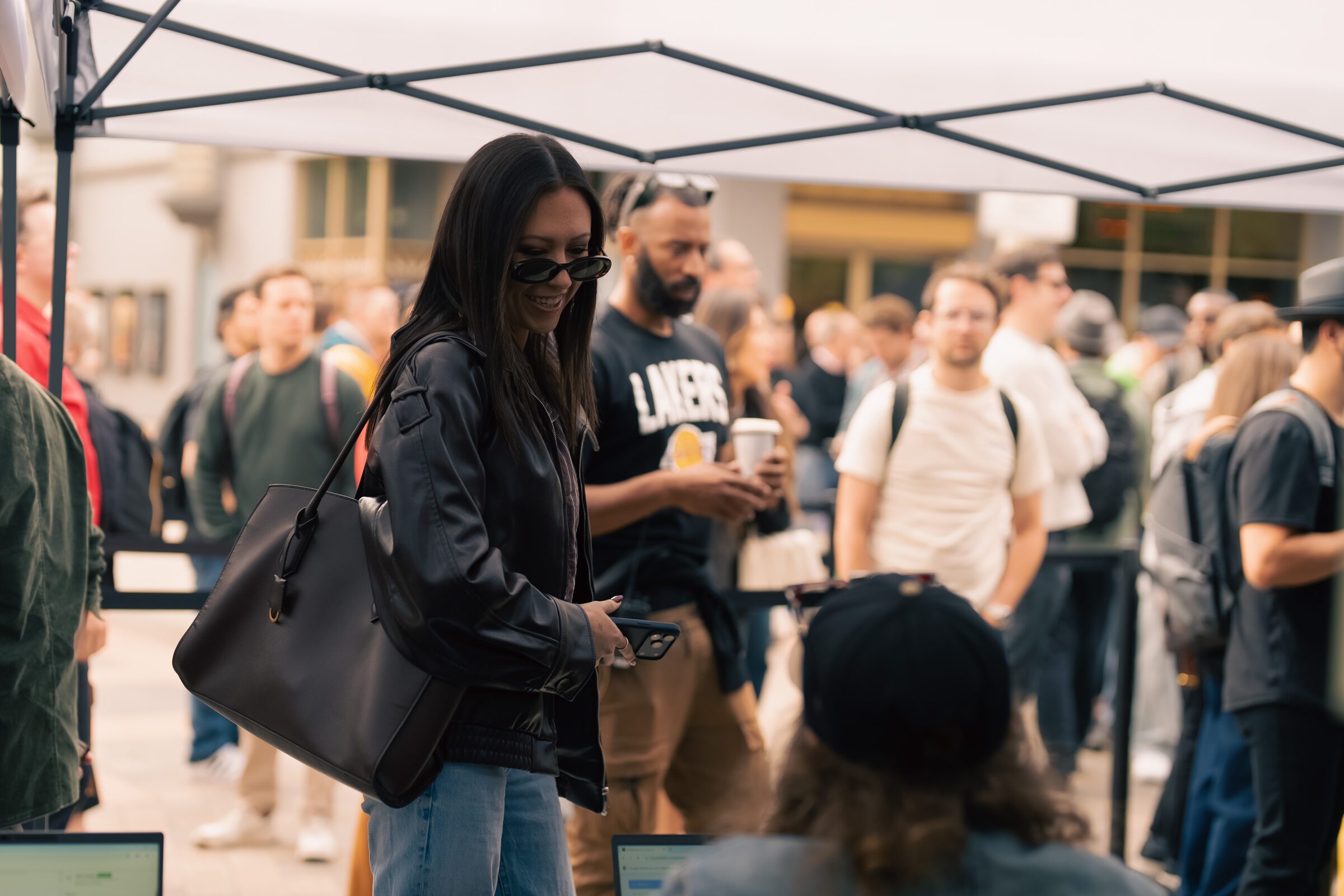 An attendee in sunglasses and a black leather jacket checks her phone at the registration tent amid a crowd.