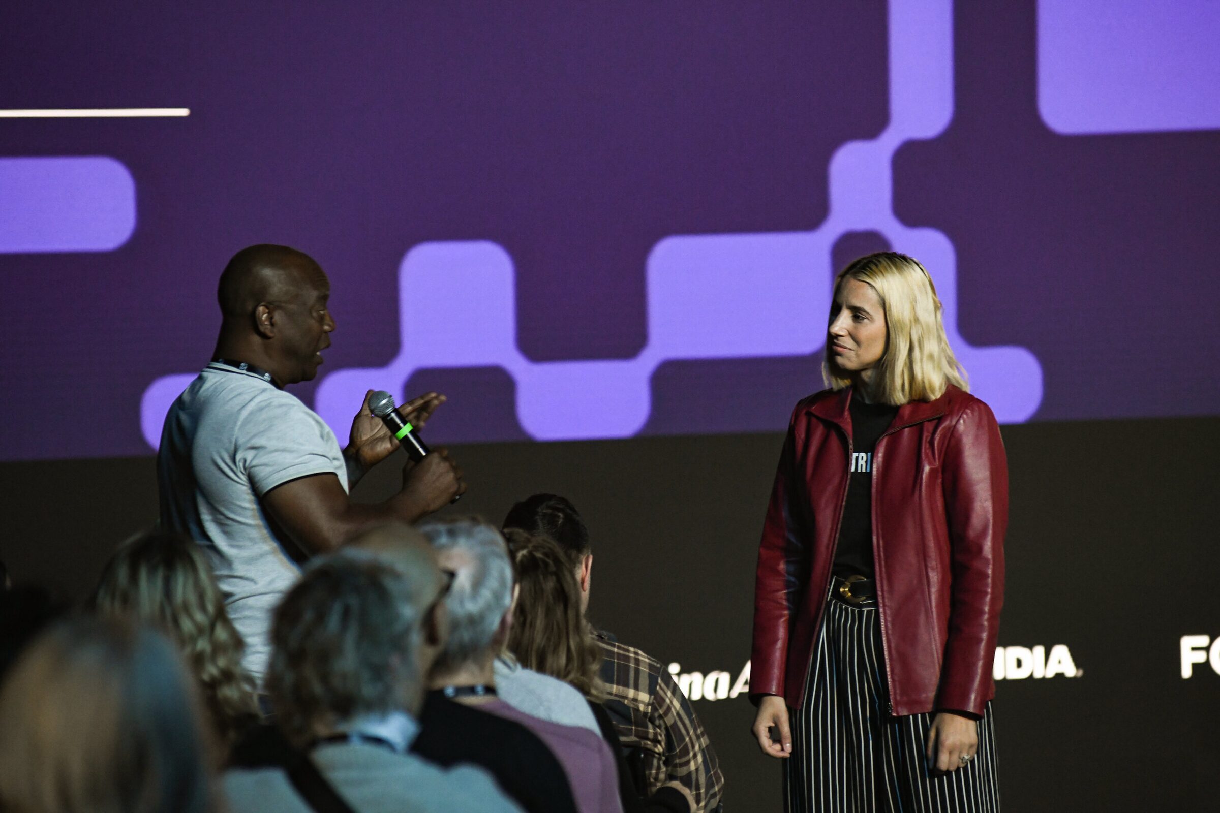 Verena Puhm, in a red leather jacket, listens as a seated attendee answers into her handheld mic.