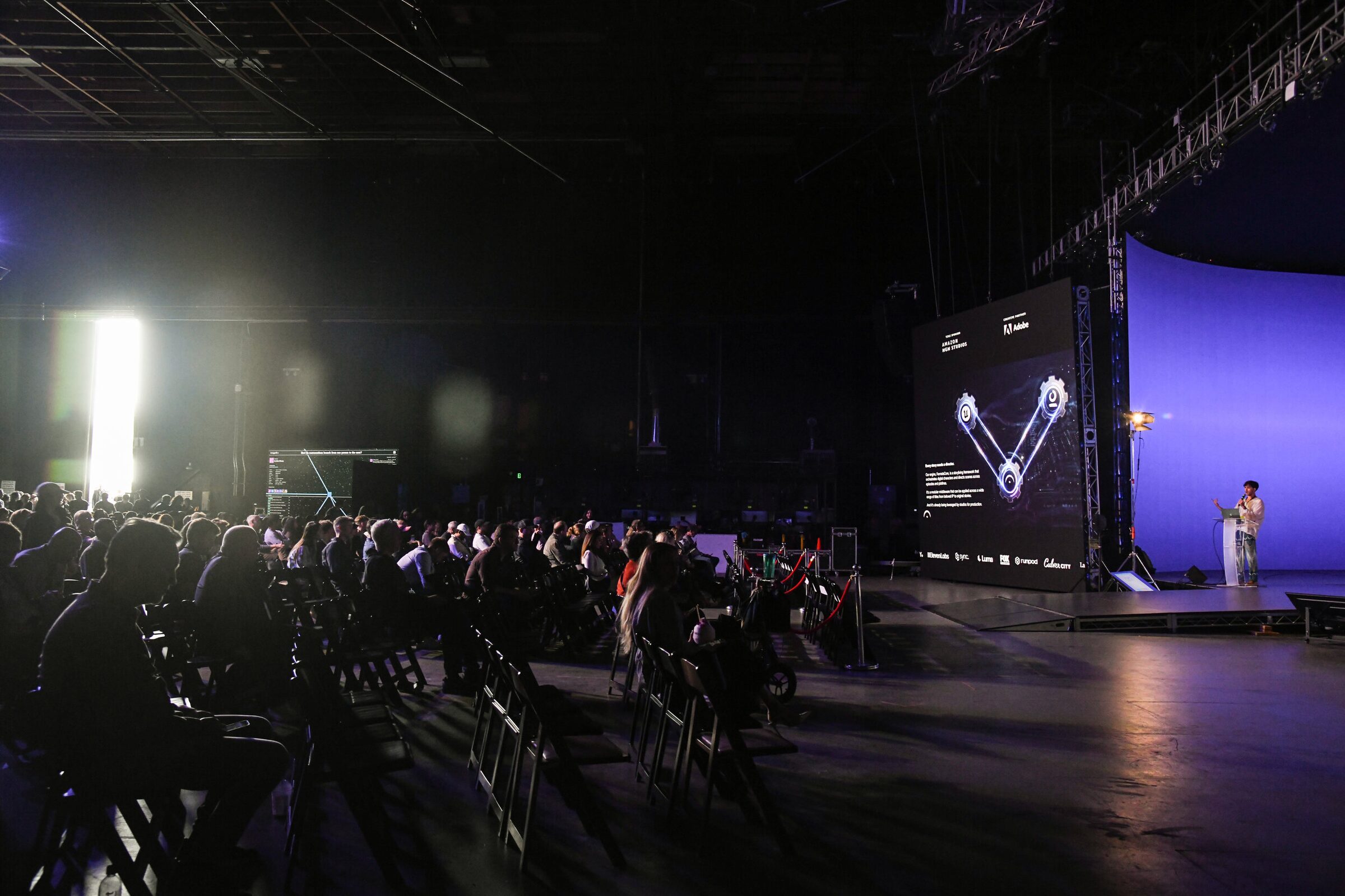 A dark Stage 15 audience faces a distant lit stage and screen, where Cameron Kostopoulos stands at a lectern.