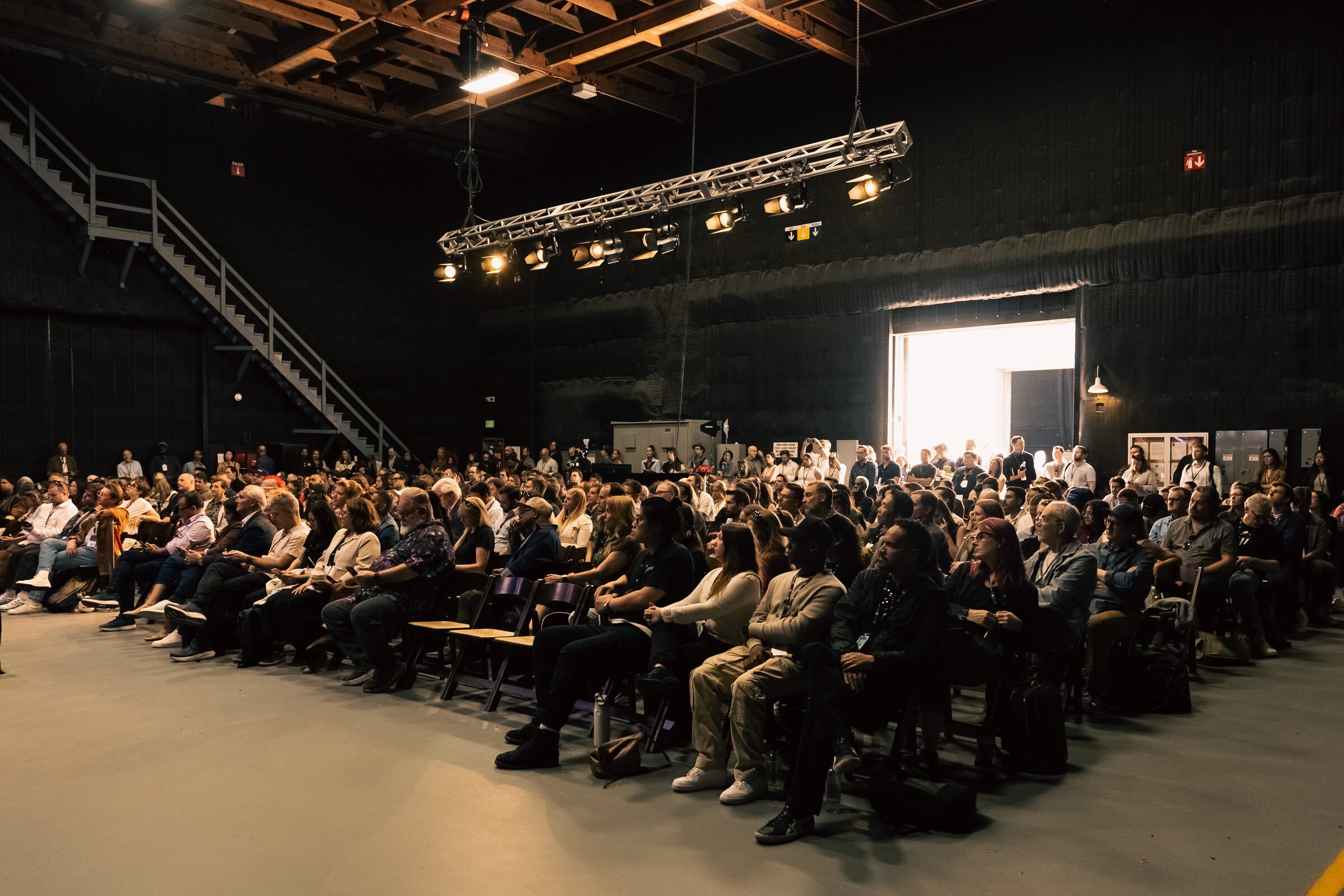 A big audience in rows faces a Google presentation in a high, beamed stage, sunlight pouring through a door.