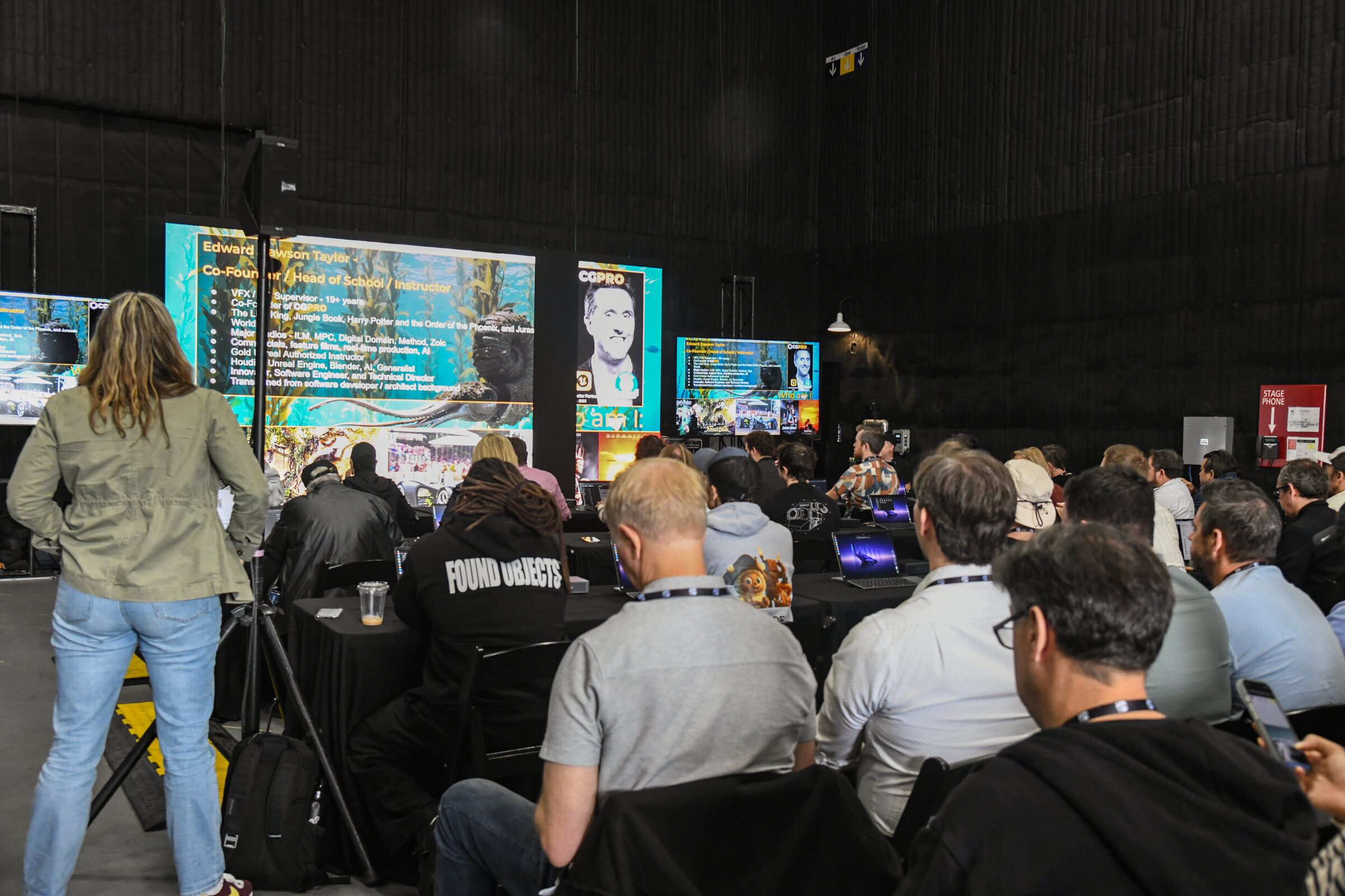 Attendees with laptops at draped tables watch CG Pro's virtual production slides on large screens in a dark stage.