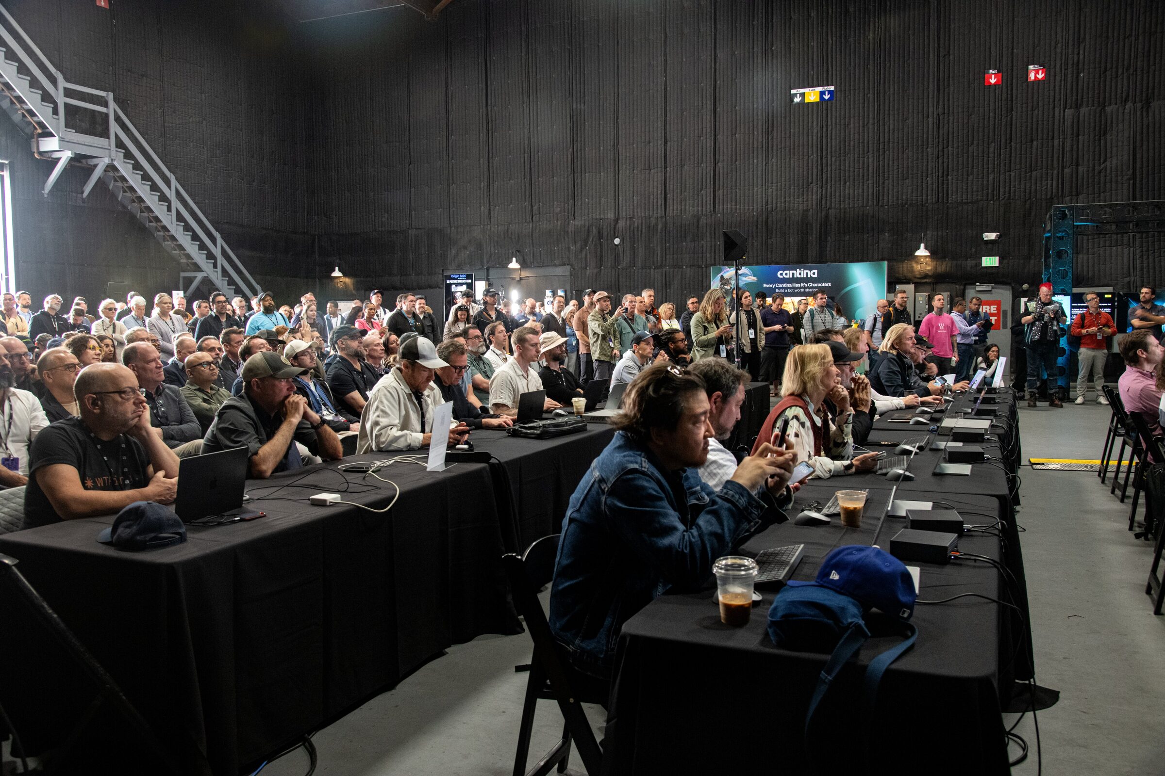 A packed workshop of attendees at long black-draped tables with laptops under a steel stairway on Stage 8.