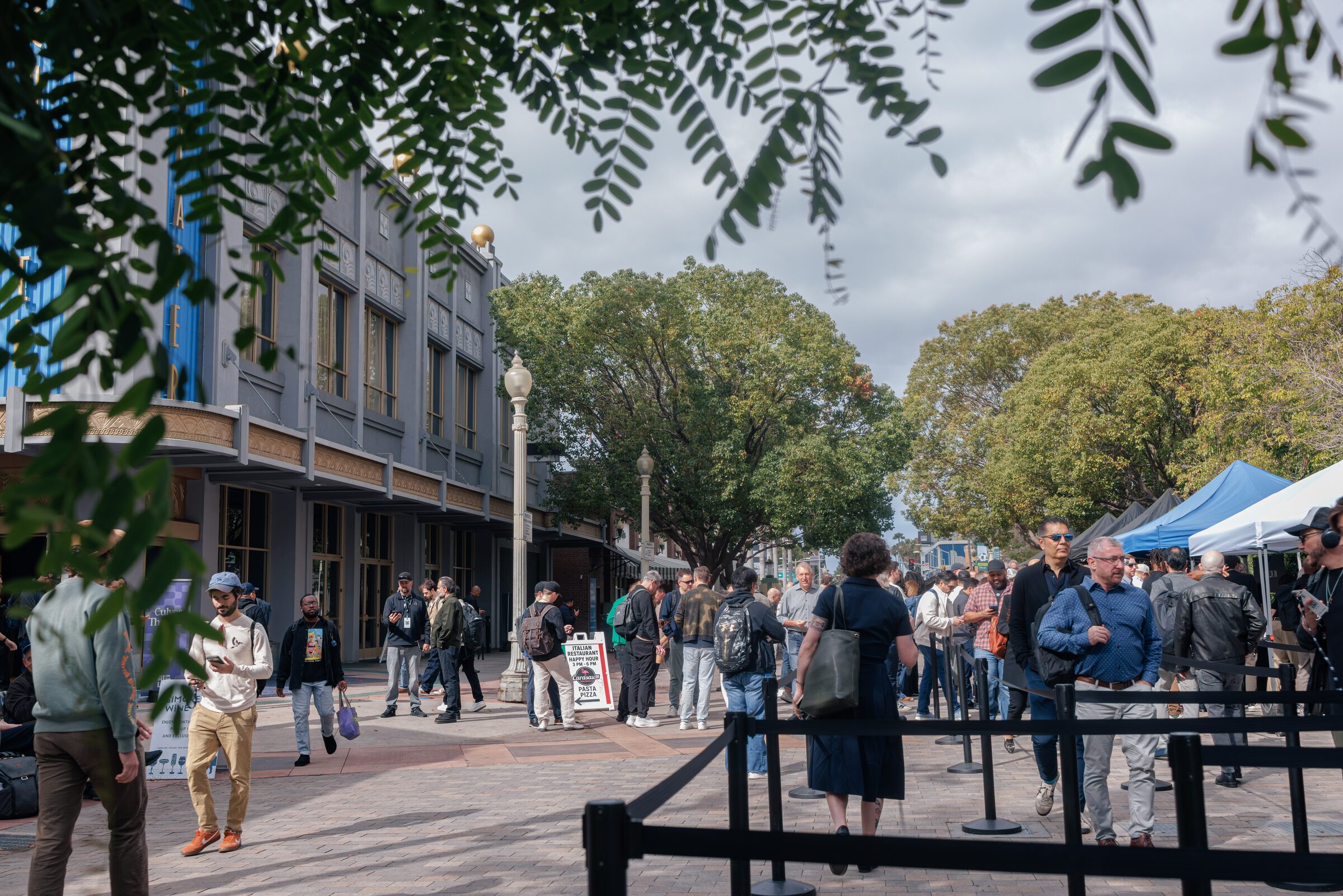 Attendees stroll and queue along stanchions on a tree-lined plaza beside a historic building and blue tents.