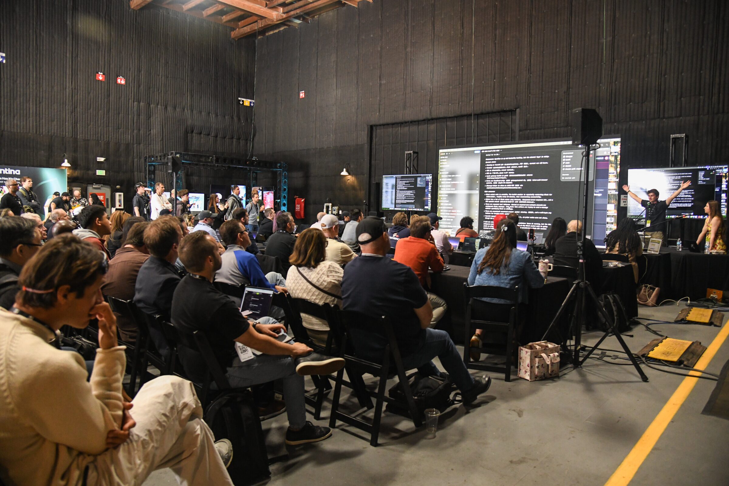 Rows of attendees with laptops face presenter screens in a dark soundstage set up for a workshop.