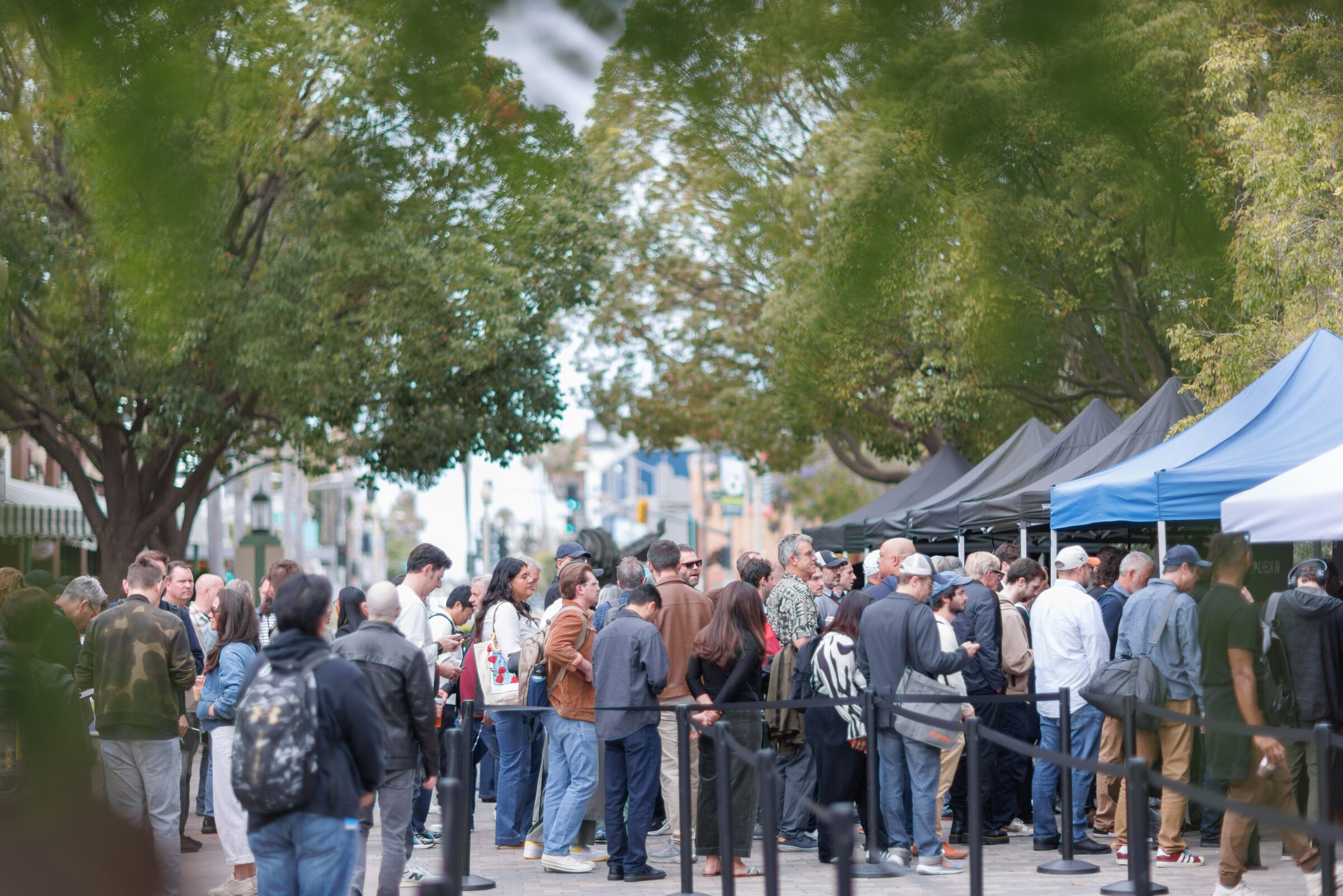A long line of attendees waits behind stanchions under leafy trees, blue registration tents along the street.
