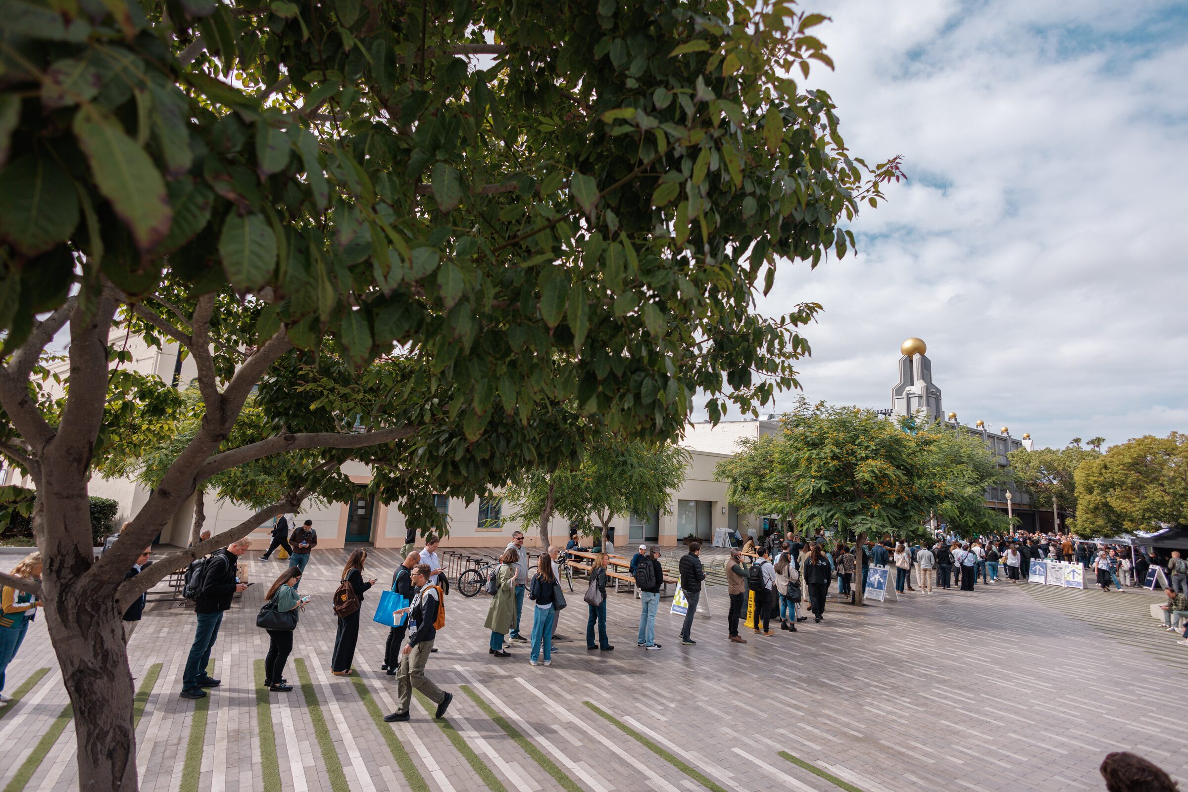 A long line of attendees snakes across a sunny, tree-shaded plaza toward a domed tower under a cloudy sky.