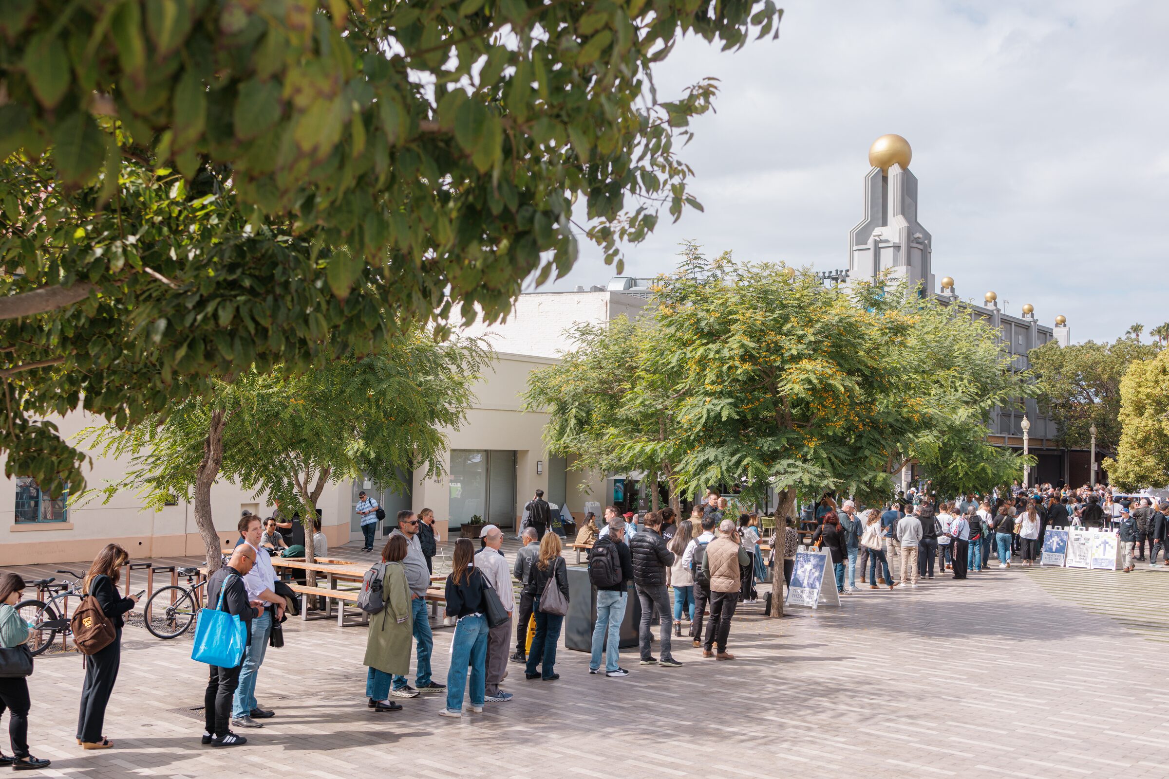 Attendees with bags queue across a paved plaza under trees, a gold-domed tower rising behind them.