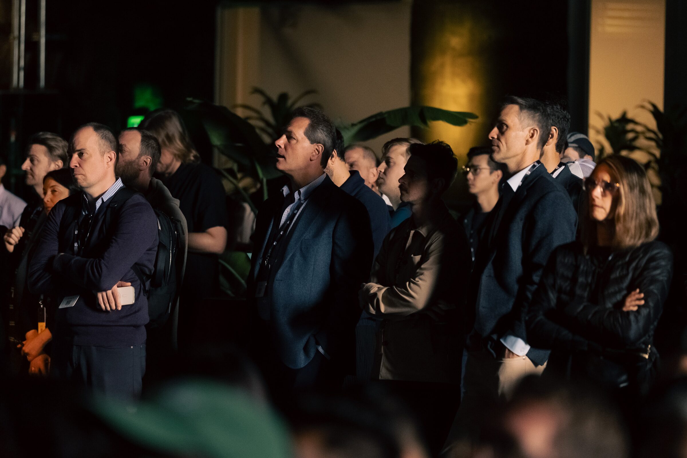Attendees in jackets stand close together and listen in a dim, plant-filled room during a private happy hour.