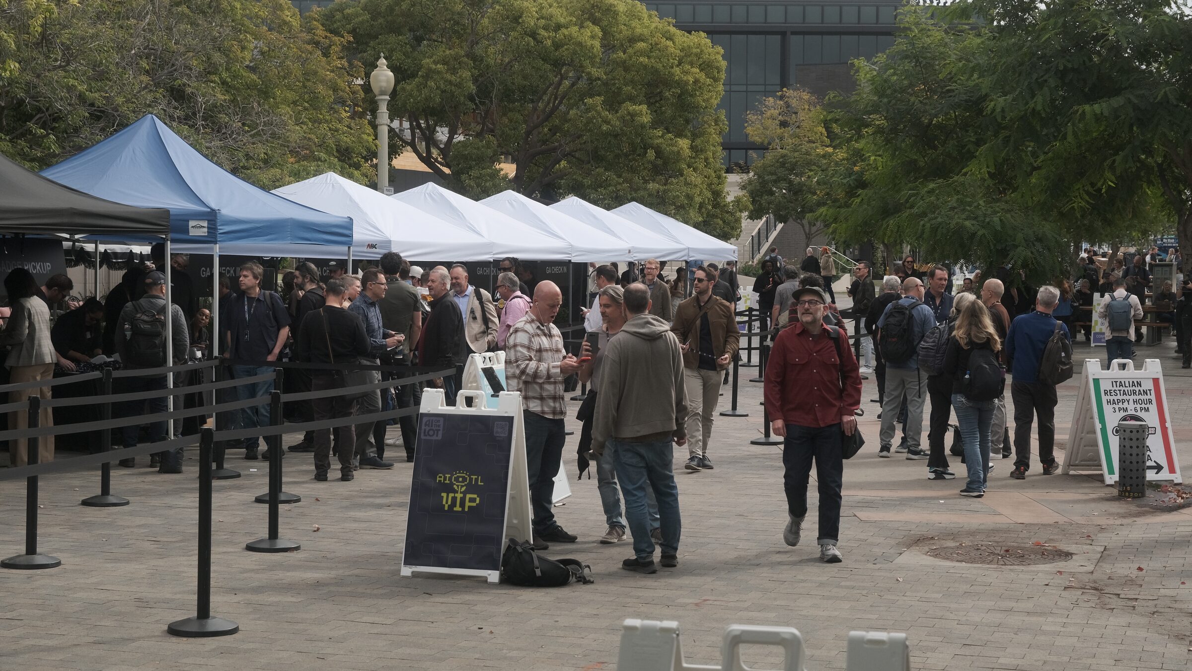 Attendees mingle and queue by stanchions and white and blue tents on a tree-lined town plaza.