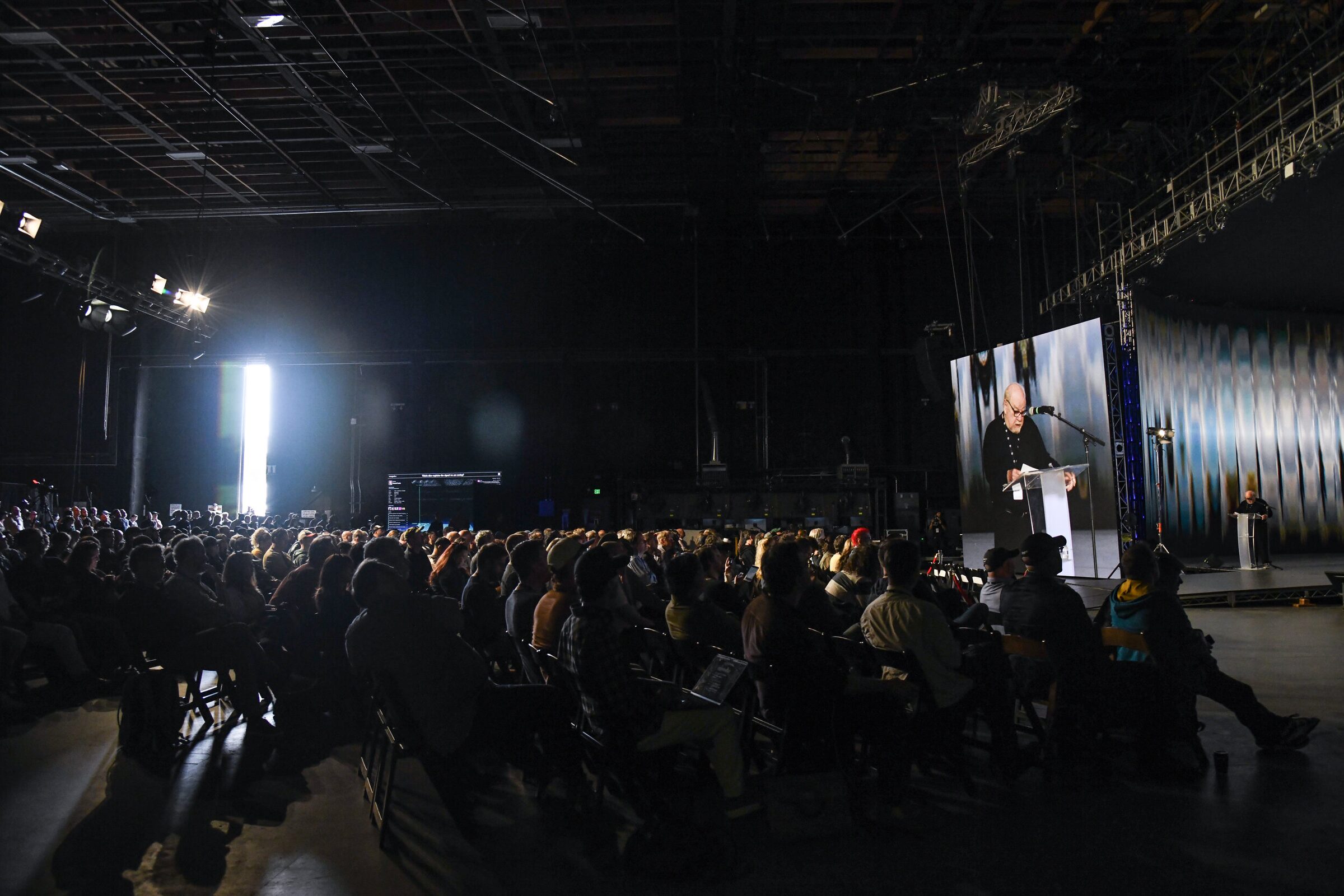 A dark, packed Stage 15 seen from the side, Paul Schrader at a lectern far off and on a big screen above.