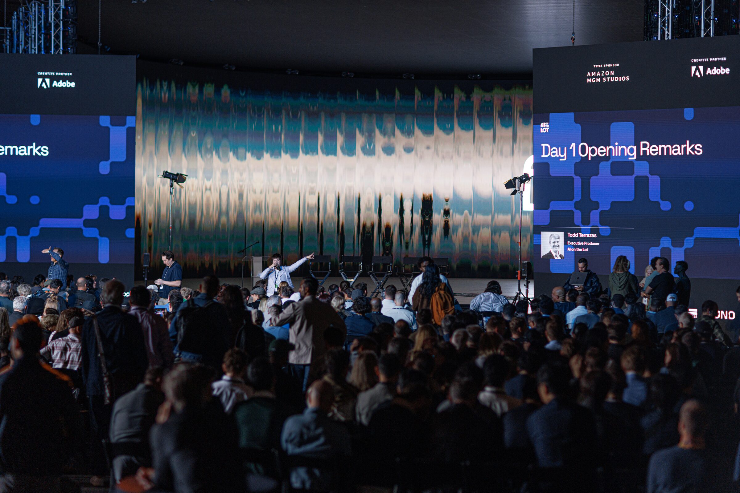A full Stage 15 crowd settles in under screens reading "Day 1 Opening Remarks," the shimmering wall behind.