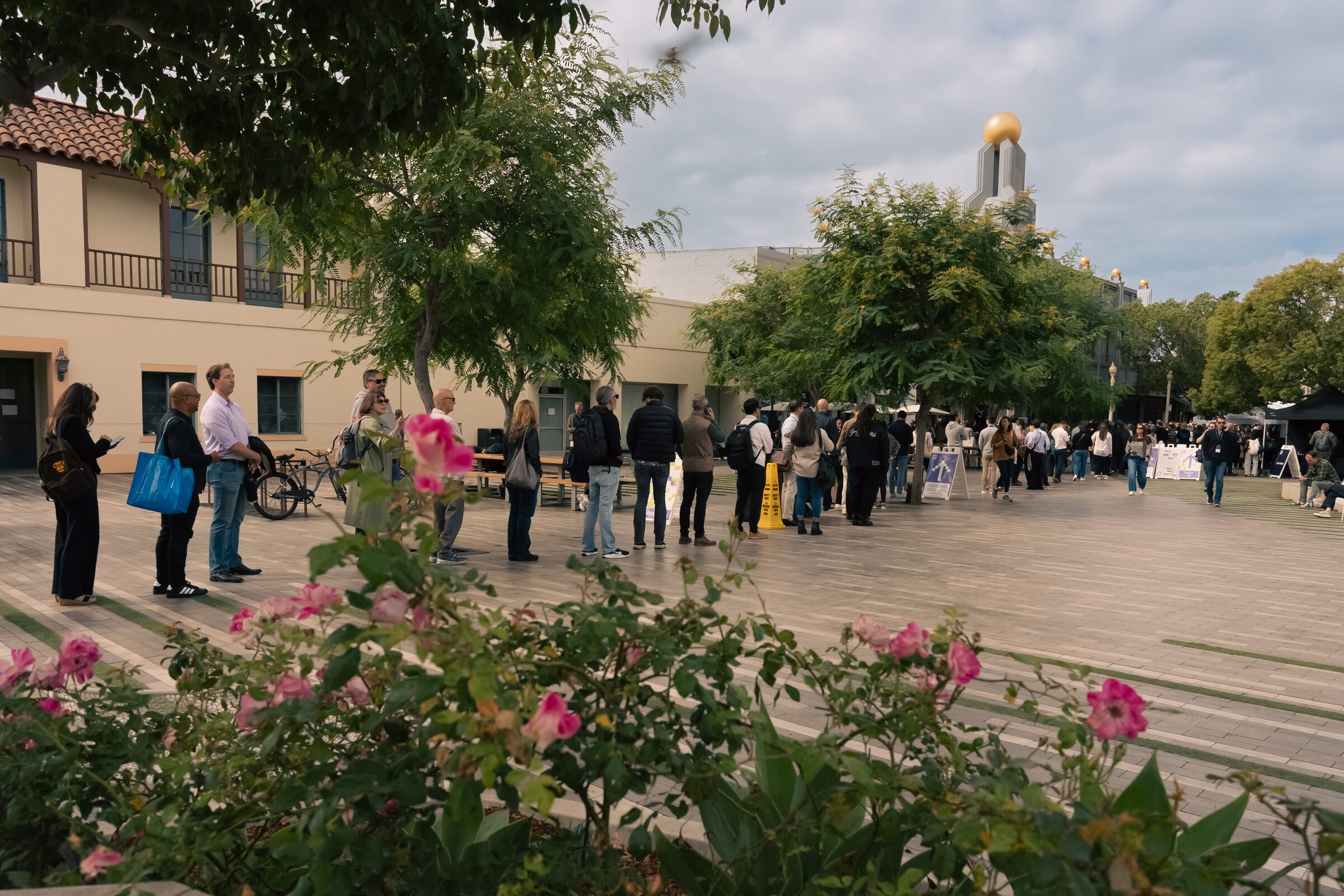 Attendees line up across a sunny plaza past pink roses, a gold-domed tower behind them.