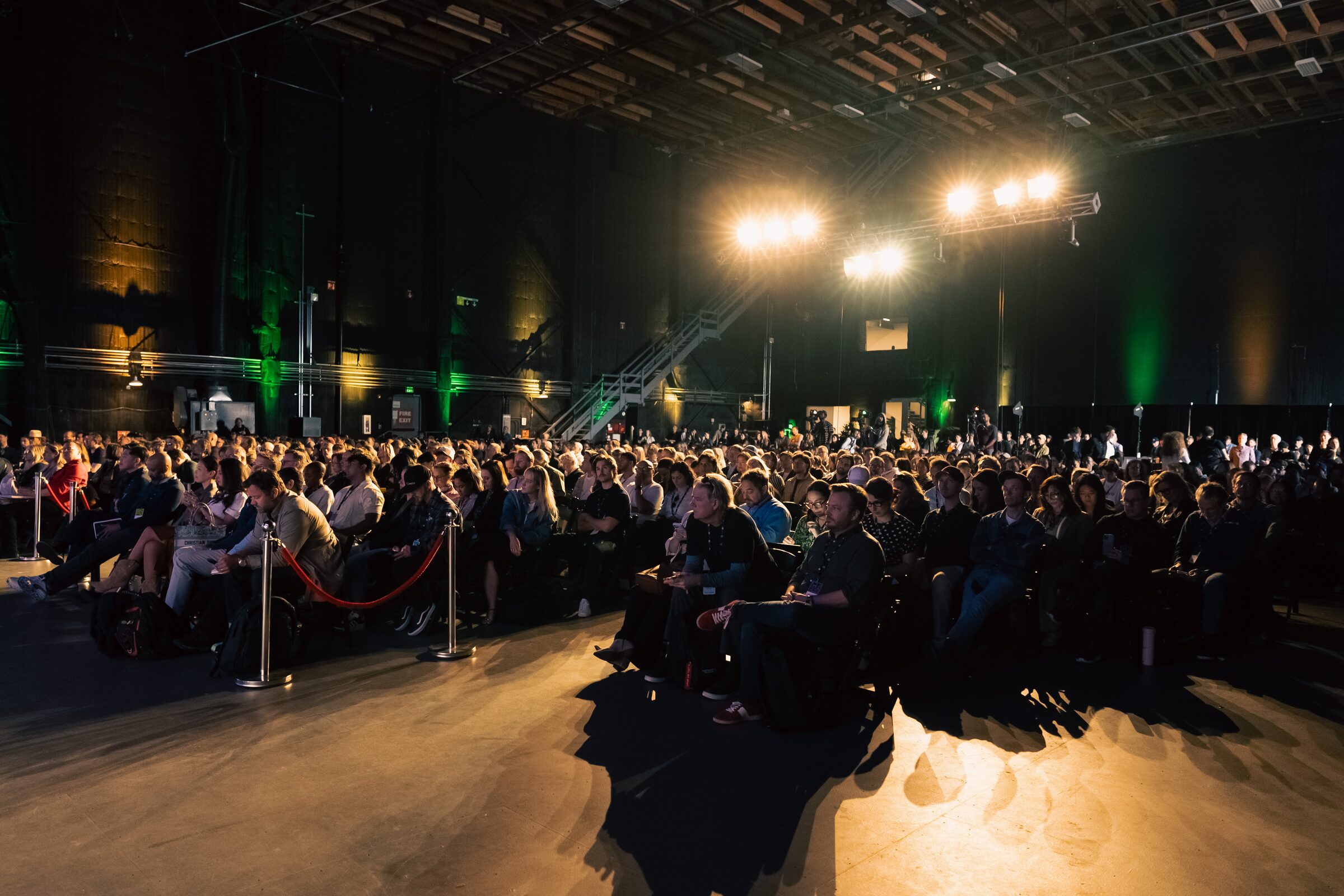 A packed audience fills rows under warm spotlights in a cavernous stage, faces turned toward the front.
