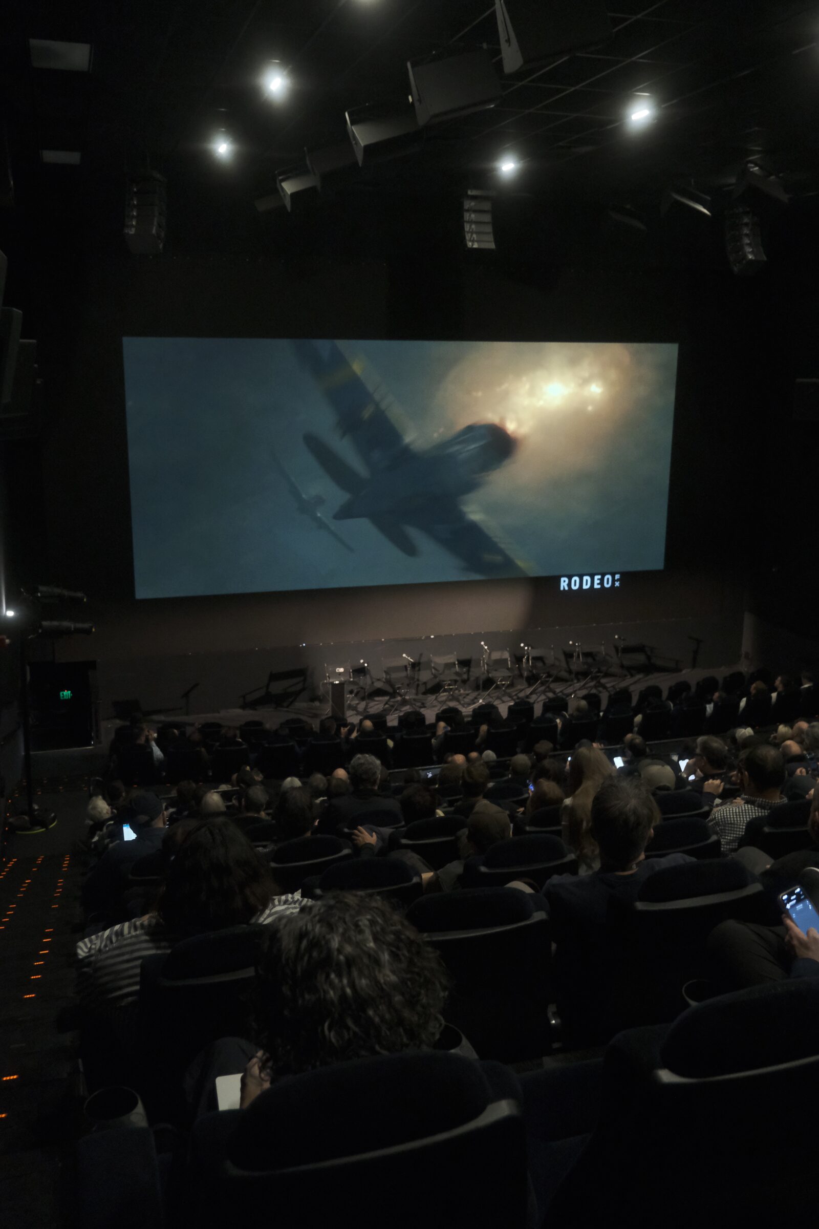 A dark theater full of attendees watches a big screen showing a fighter plane diving through smoke.