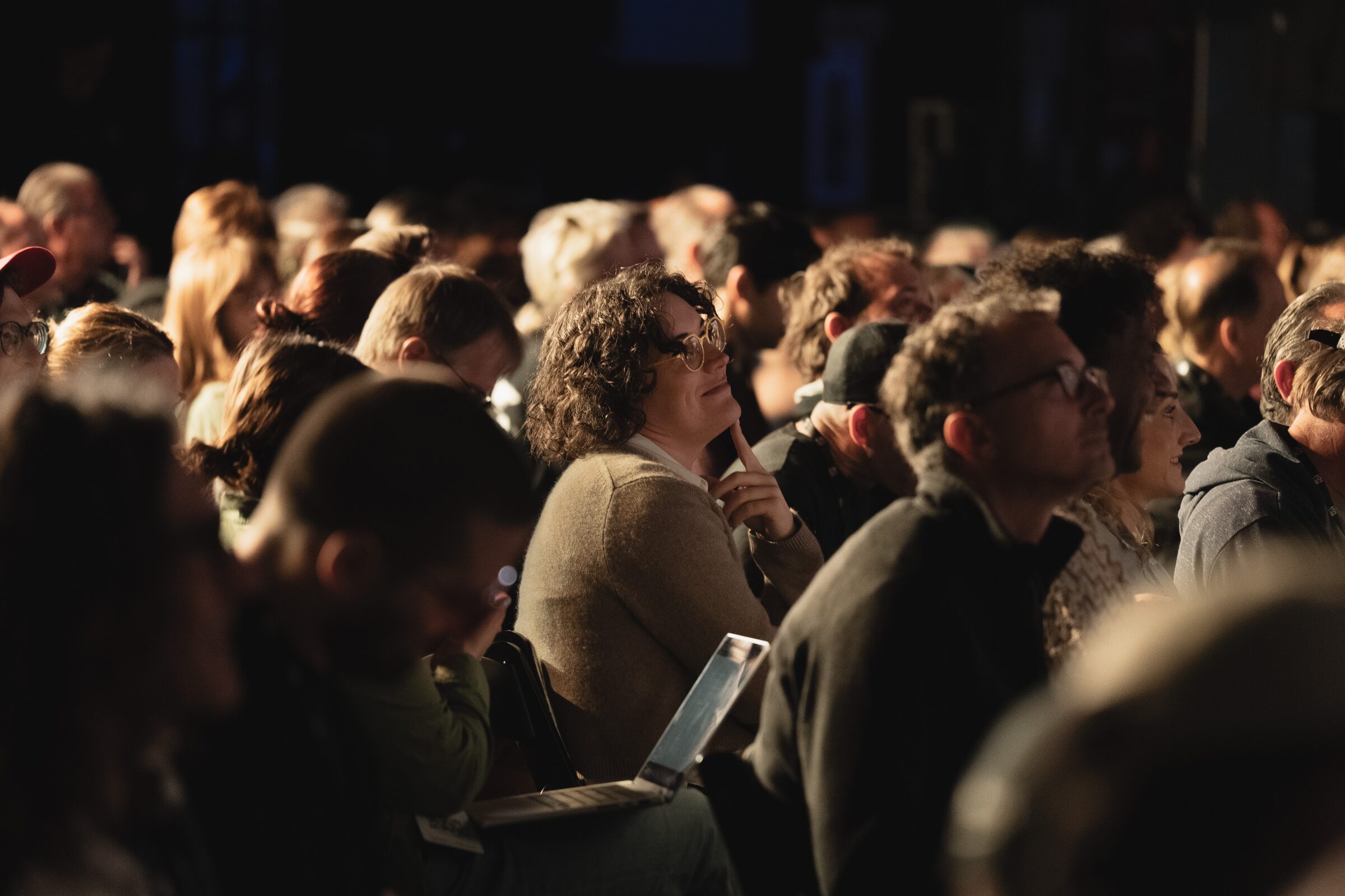 Close-up of a rapt audience in the dark, one attendee with a laptop open, all looking up toward the stage.