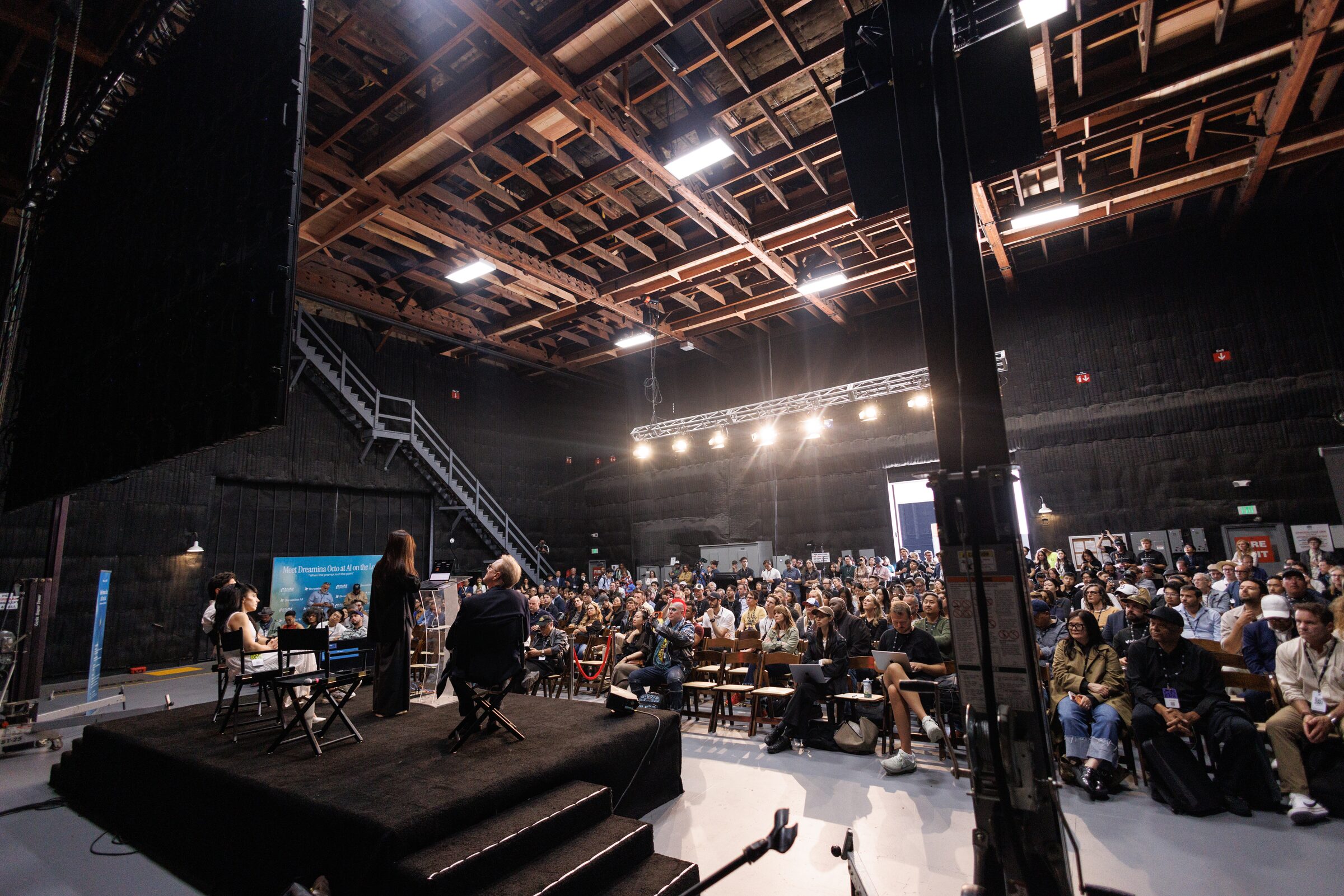 A full, sunlit workshop stage audience faces a small dais where presenters stand by a product slide.