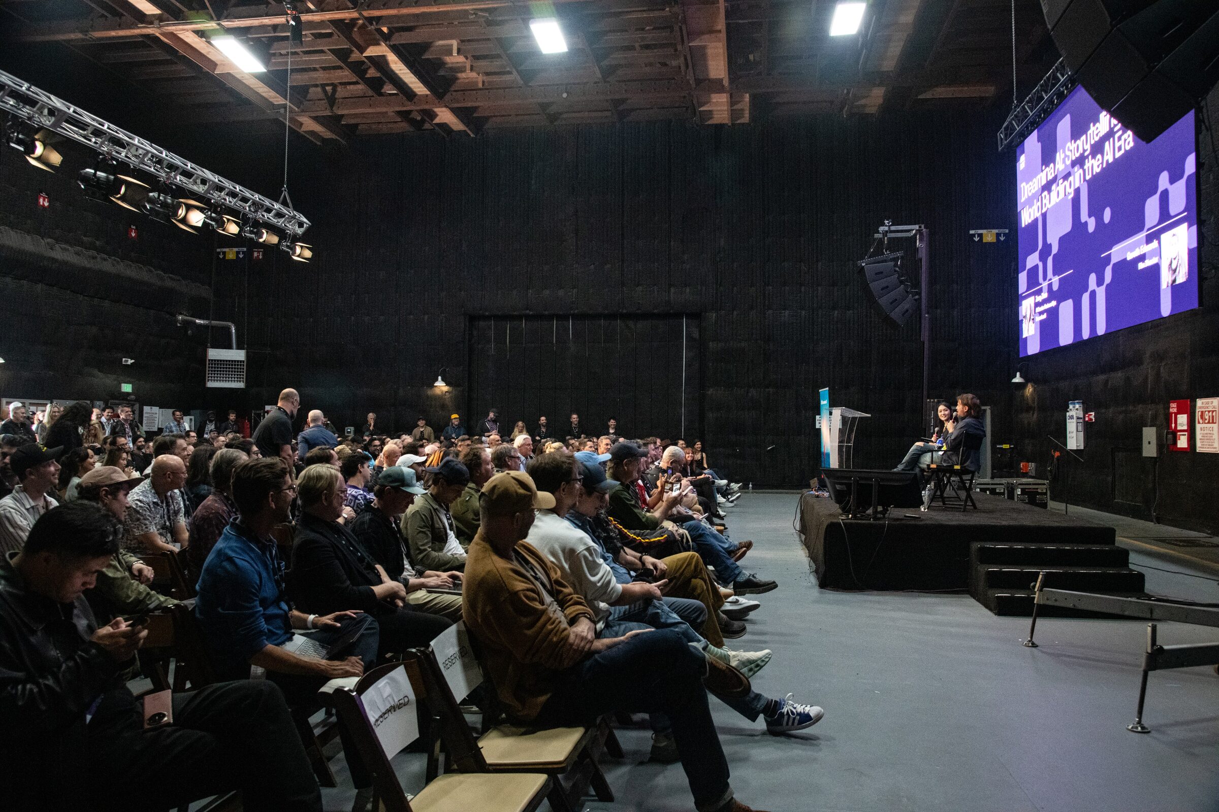 A wide view of a soundstage audience in rows facing a small stage where Gareth Edwards sits beside a big screen.