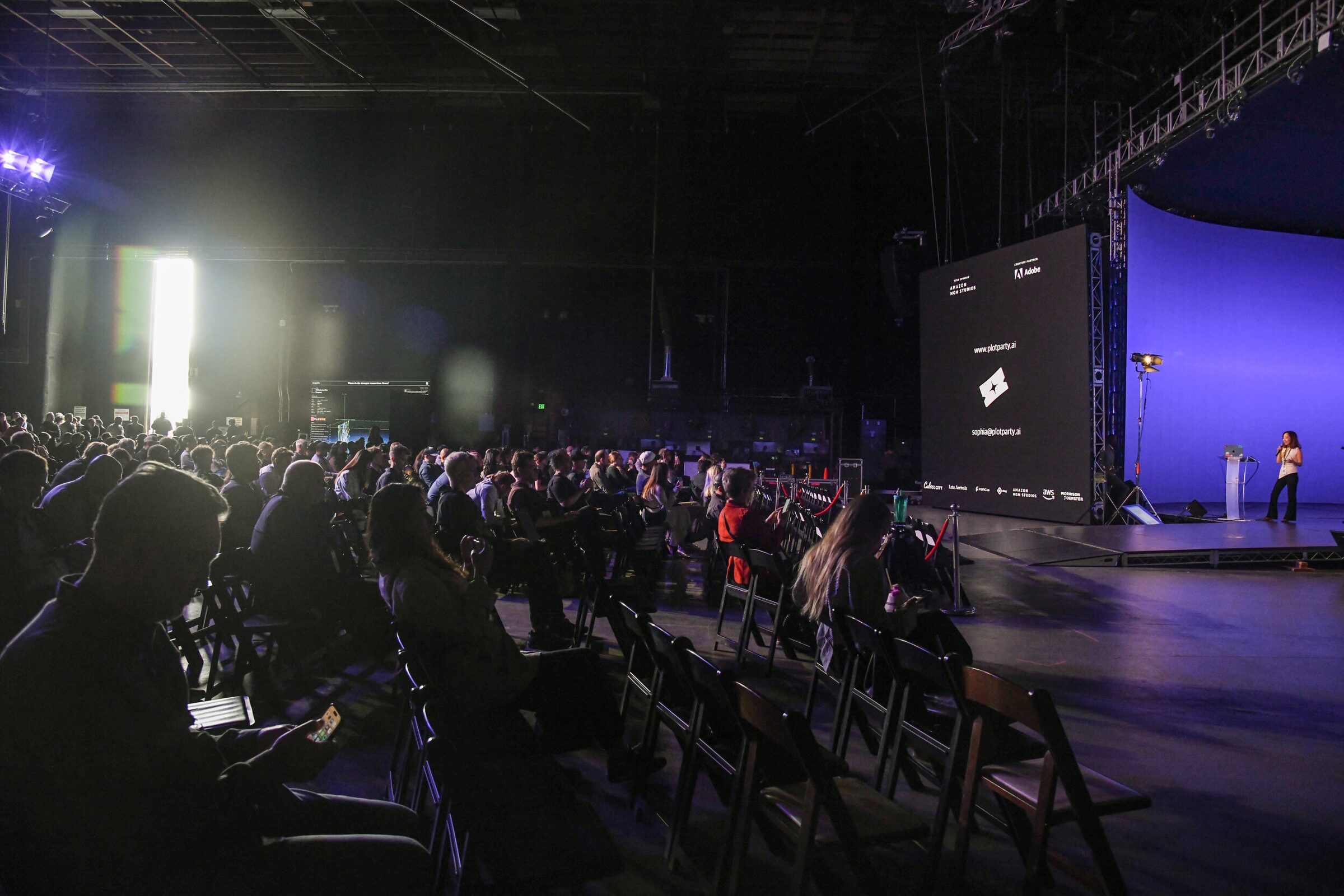 A dark, crowded Stage 15 audience watches a speaker at a lectern on the far blue stage beside a big screen.