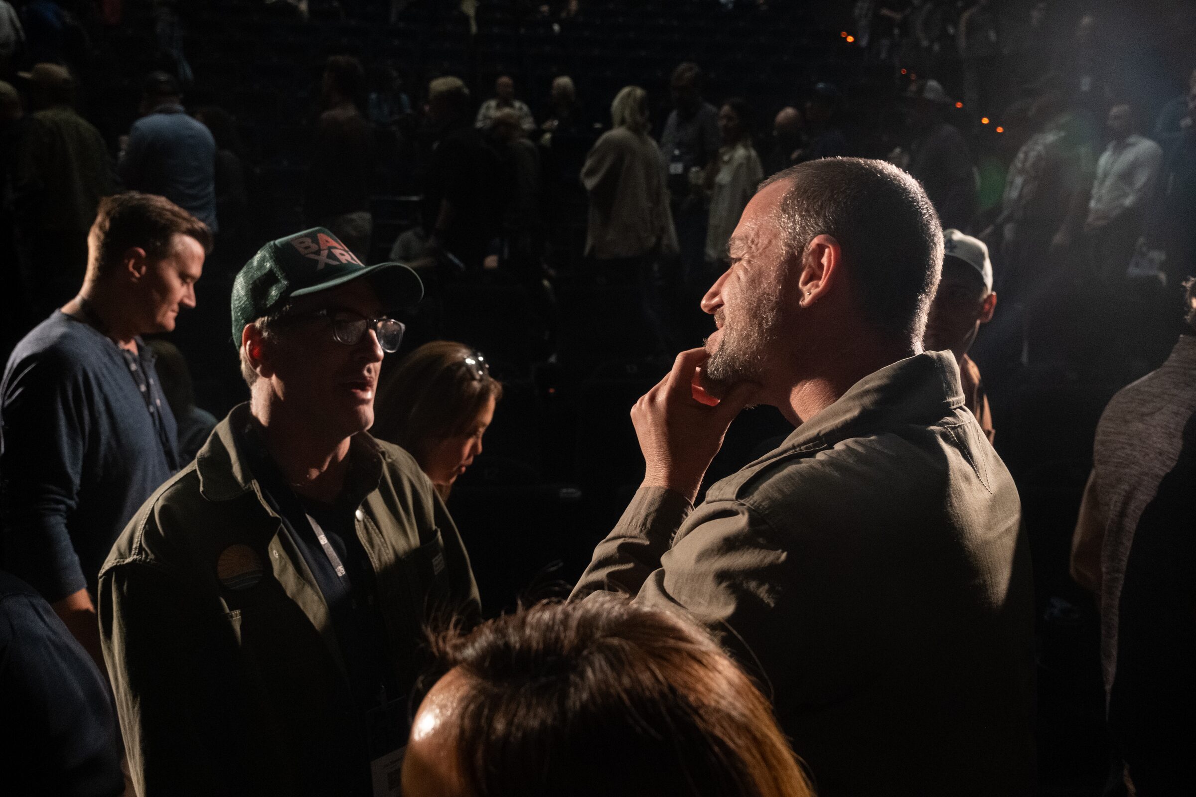 A man in a cap and glasses asks Bryn Mooser a question as Mooser listens, hand on his chin, in a dark crowd.