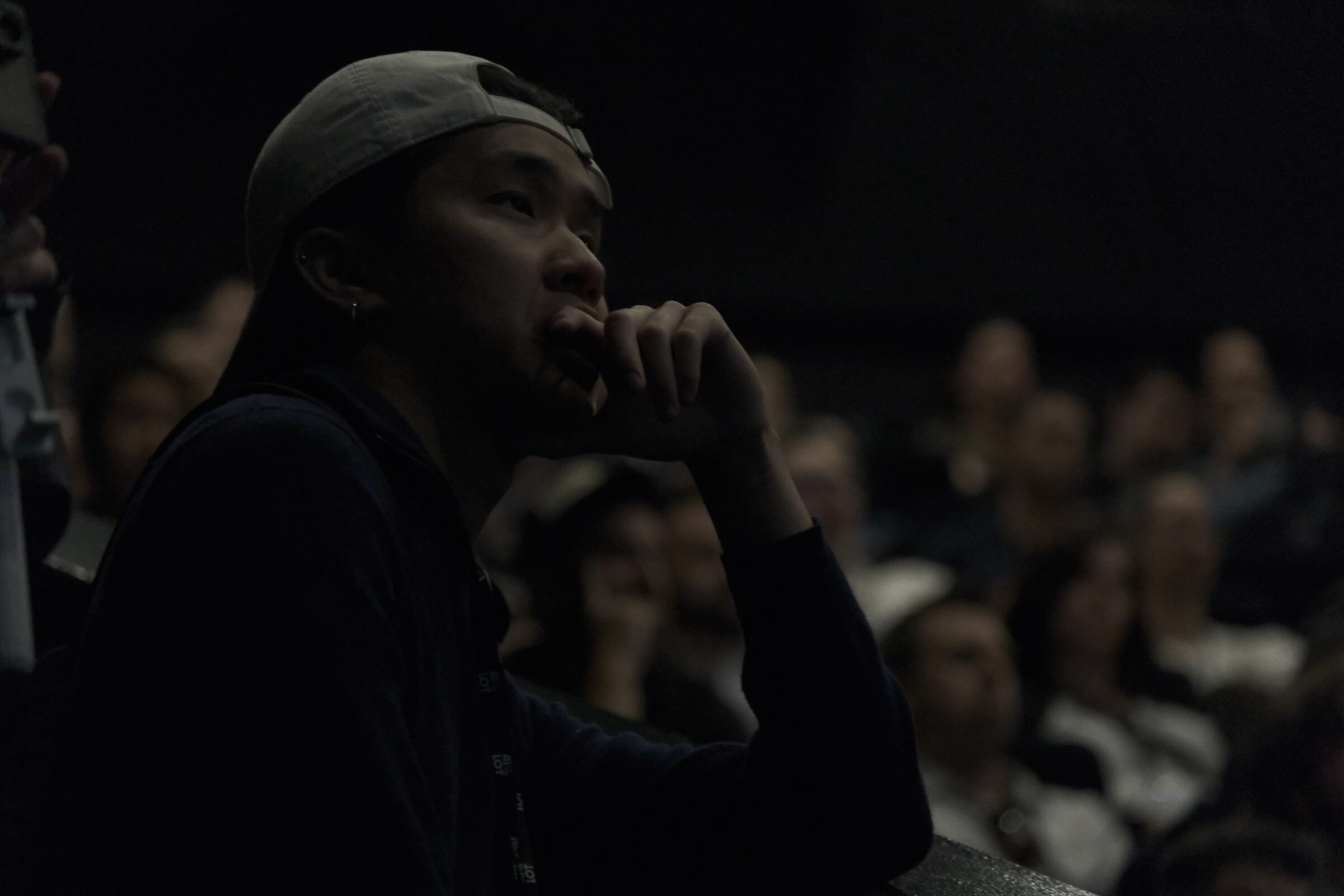 A young man in a backwards cap rests his chin on his hand, deep in thought, in a dark theater audience.
