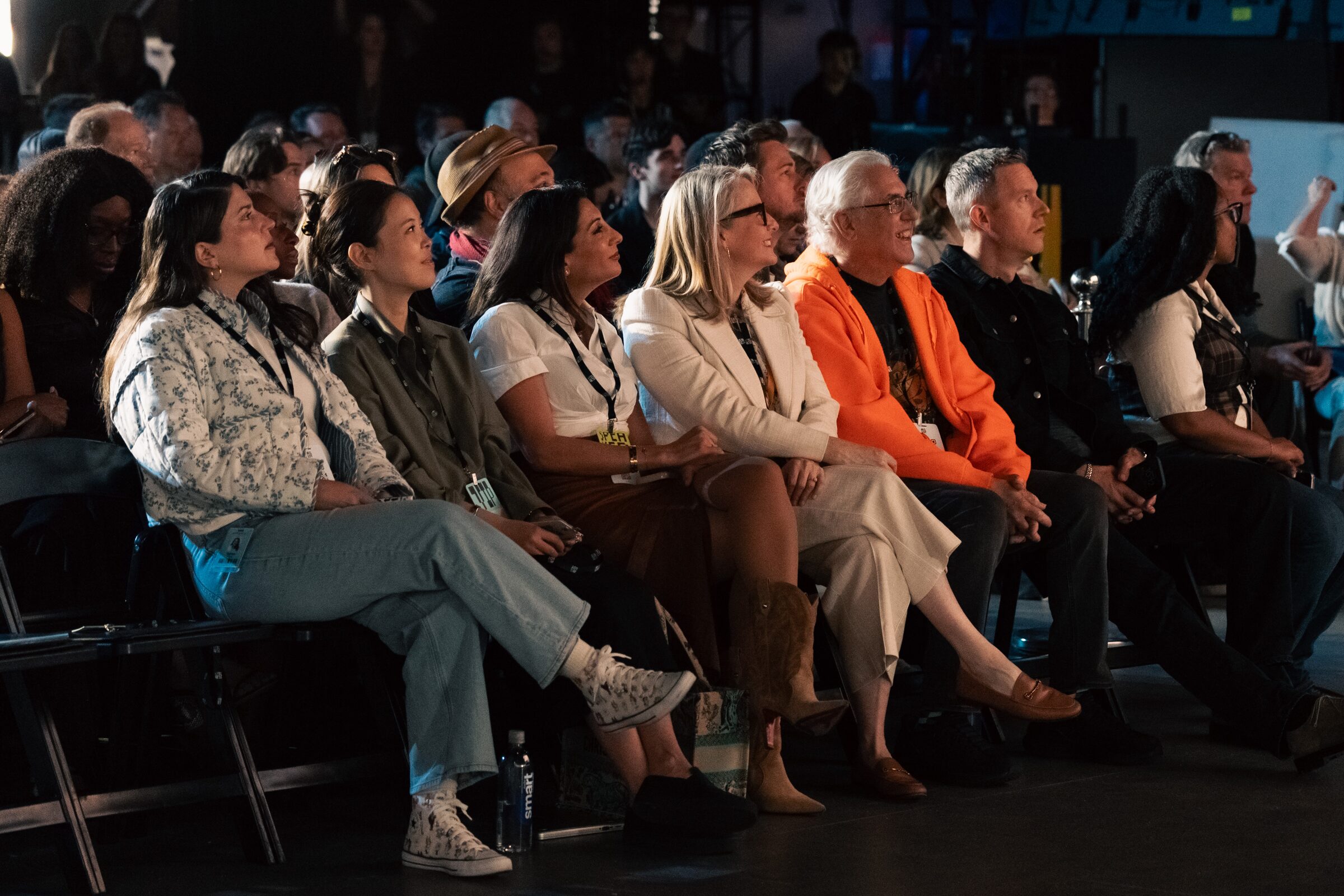 A front row of attendees in bright jackets leans in and smiles, watching the stage from their seats.