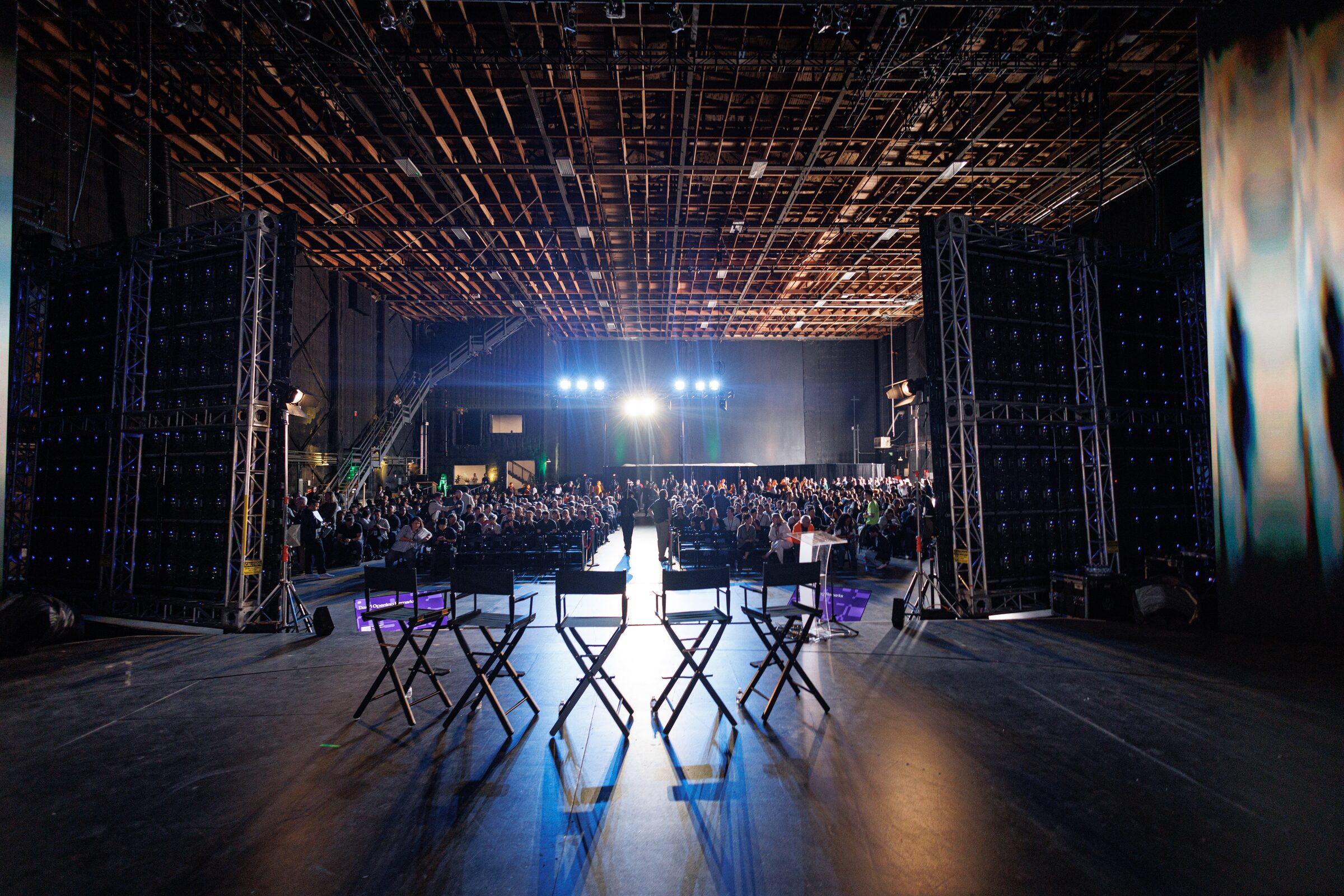 Empty director's chairs on stage face a huge crowd, the lighting grid and bright lights hanging overhead.