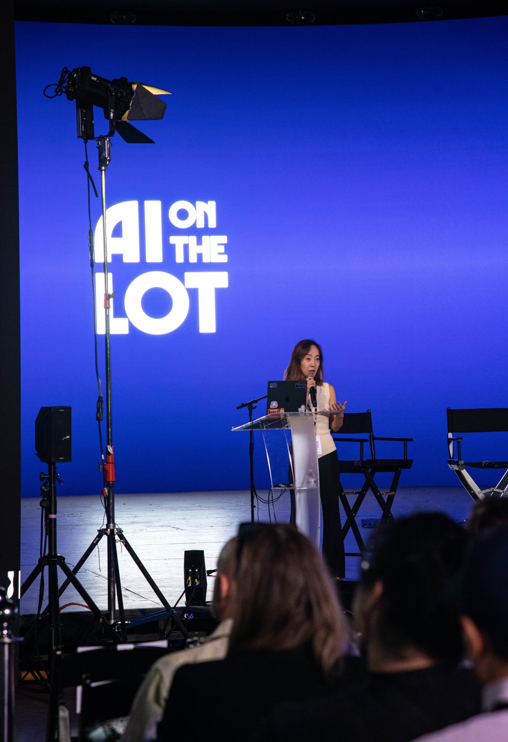 Sophia Xing at a podium on a blue stage, dwarfed by the AI on the Lot logo and a studio light on a stand.