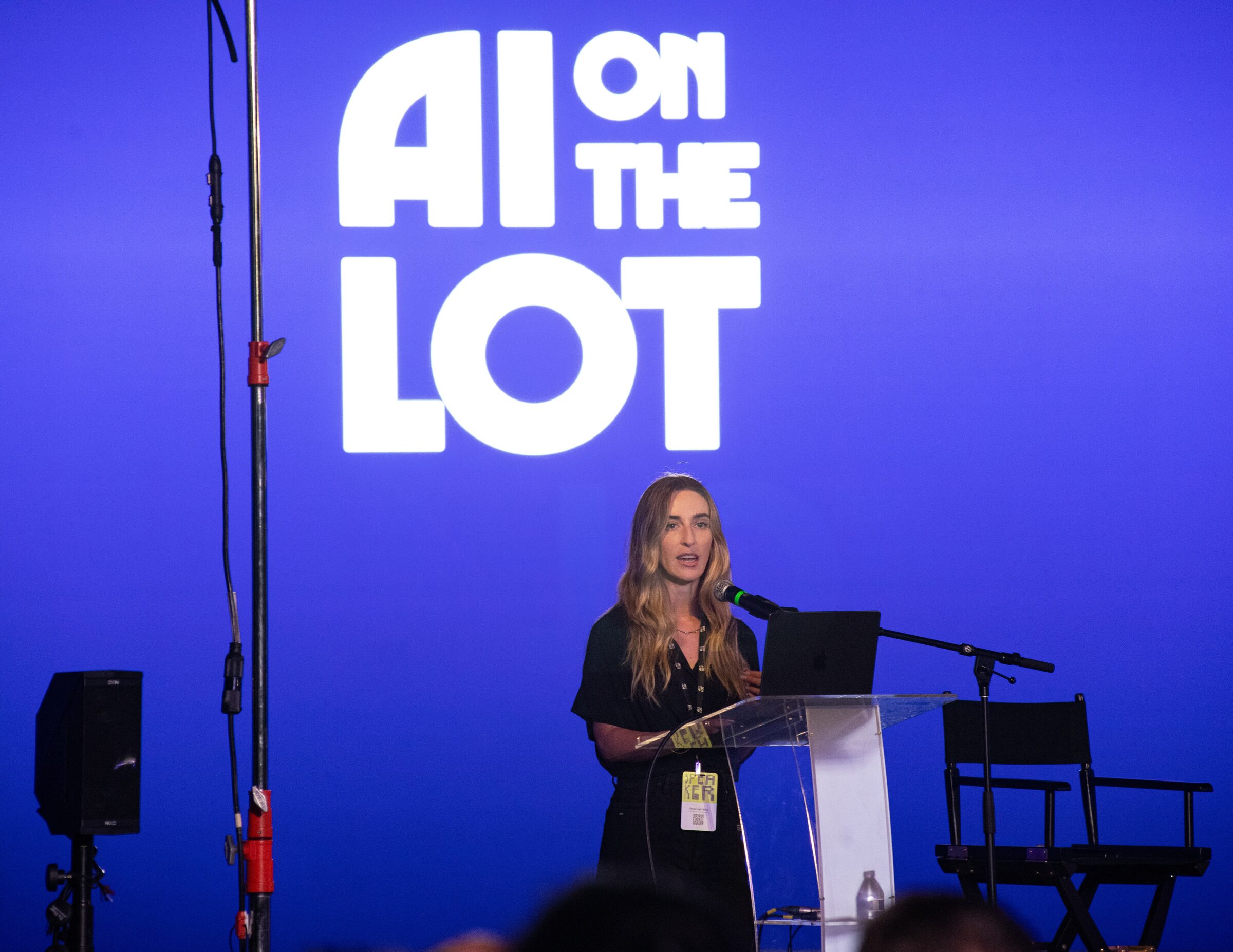 Savannah Niles speaks at a clear lectern, the white AI on the Lot logo lit on the blue wall above her.