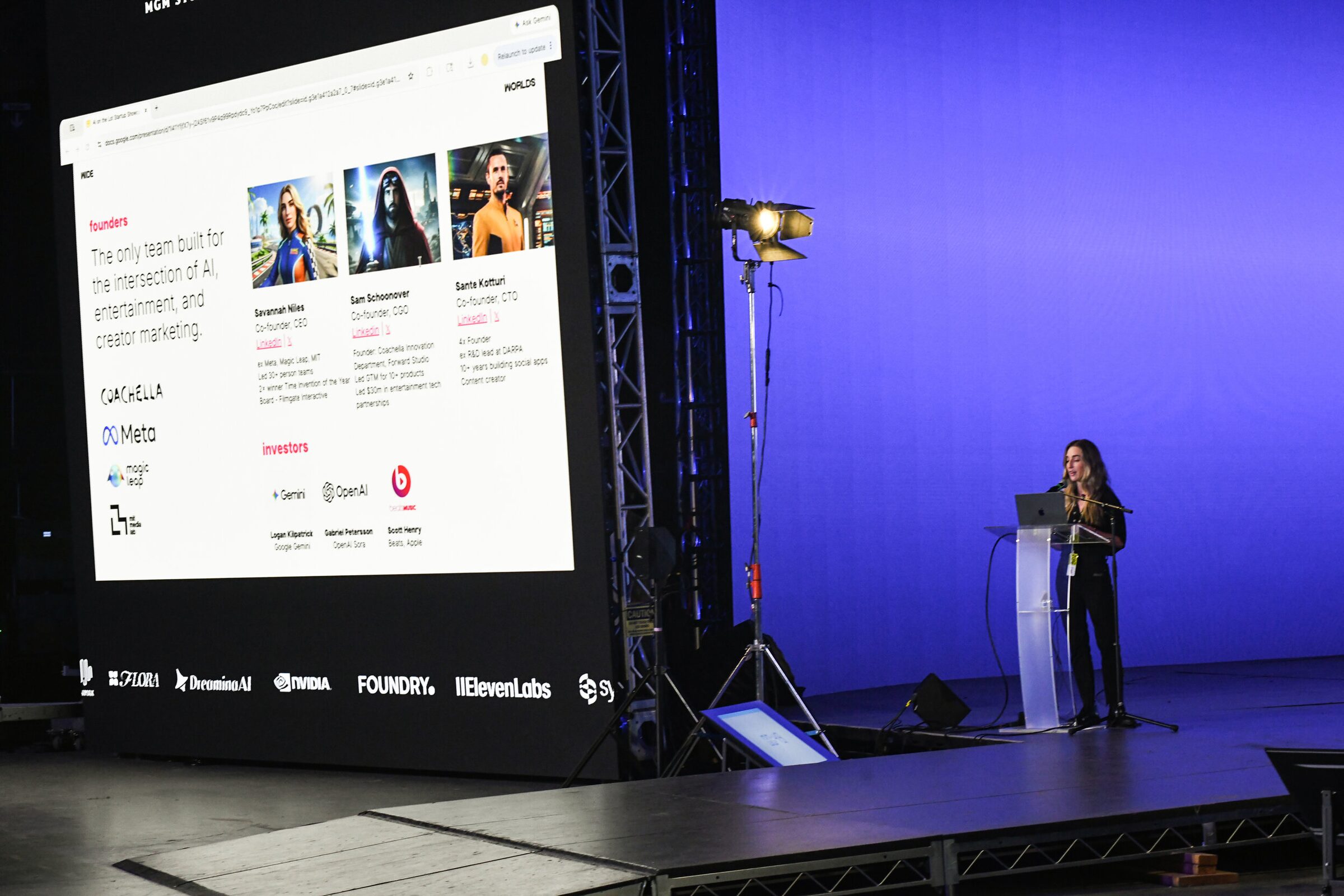 Savannah Niles at a lectern on the blue stage beside a big screen showing her team, partners and investors.