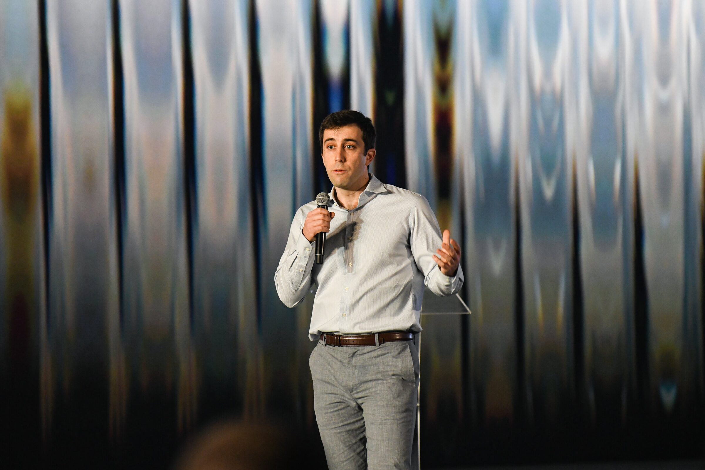 Mike Gioia speaks into a handheld mic in front of a shimmering blue and silver wall projection.