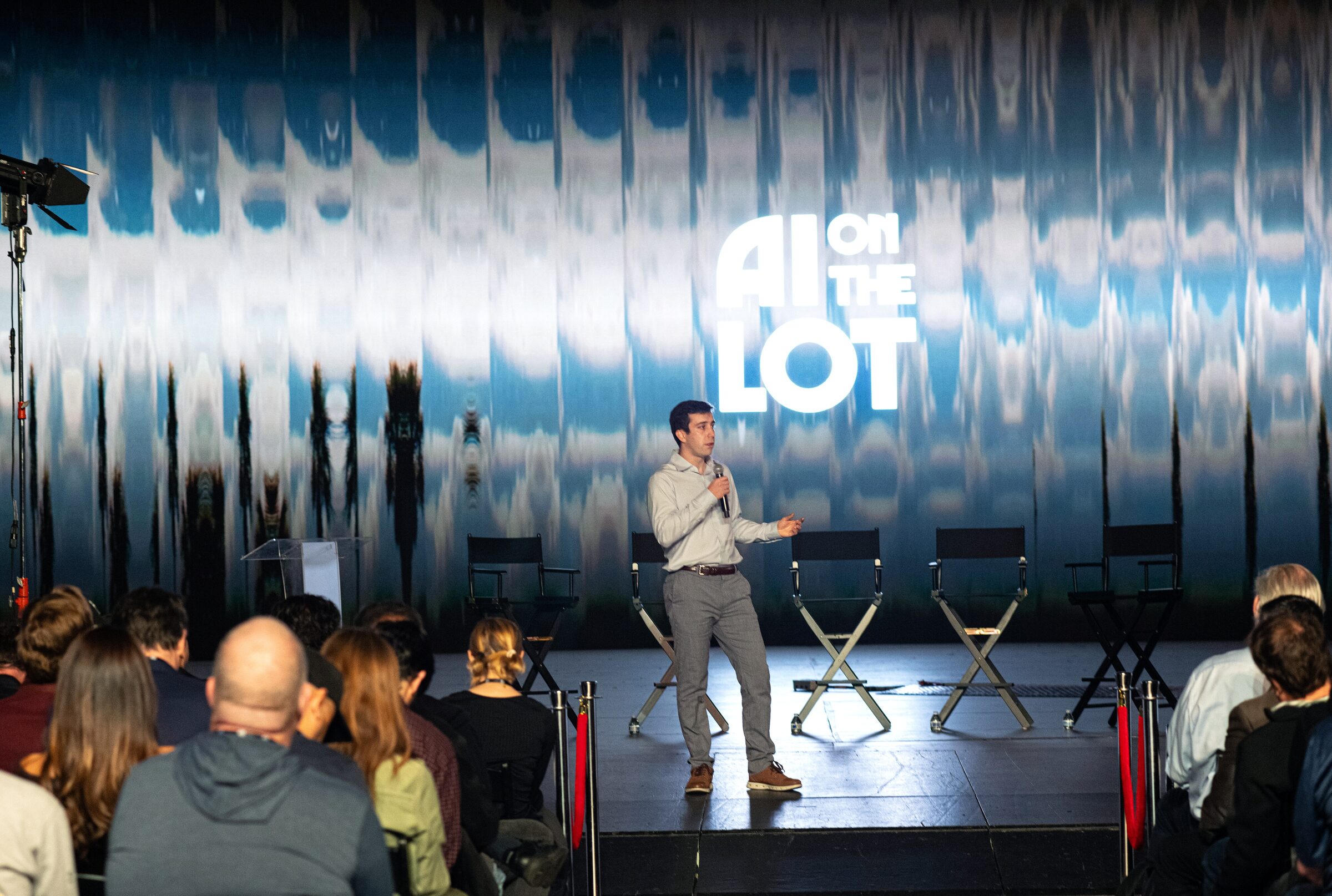 Mike Gioia on stage beside a row of empty director's chairs, seen from behind the front rows of the audience.