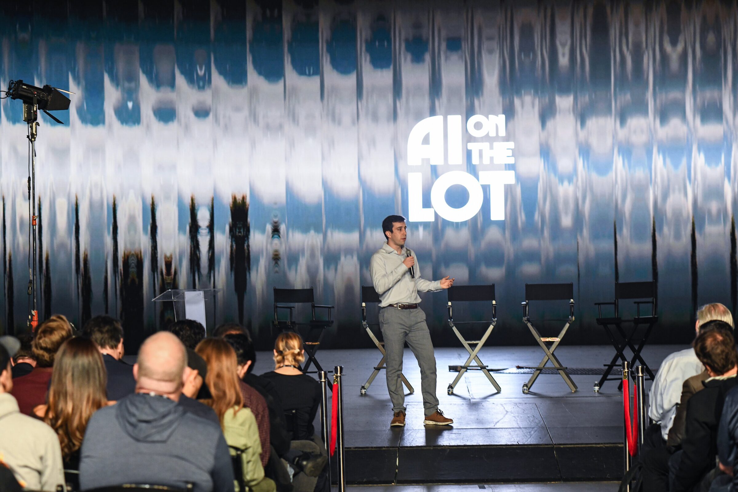 Mike Gioia stands on stage by empty director's chairs, AI on the Lot logo on the shimmering wall, crowd in front.