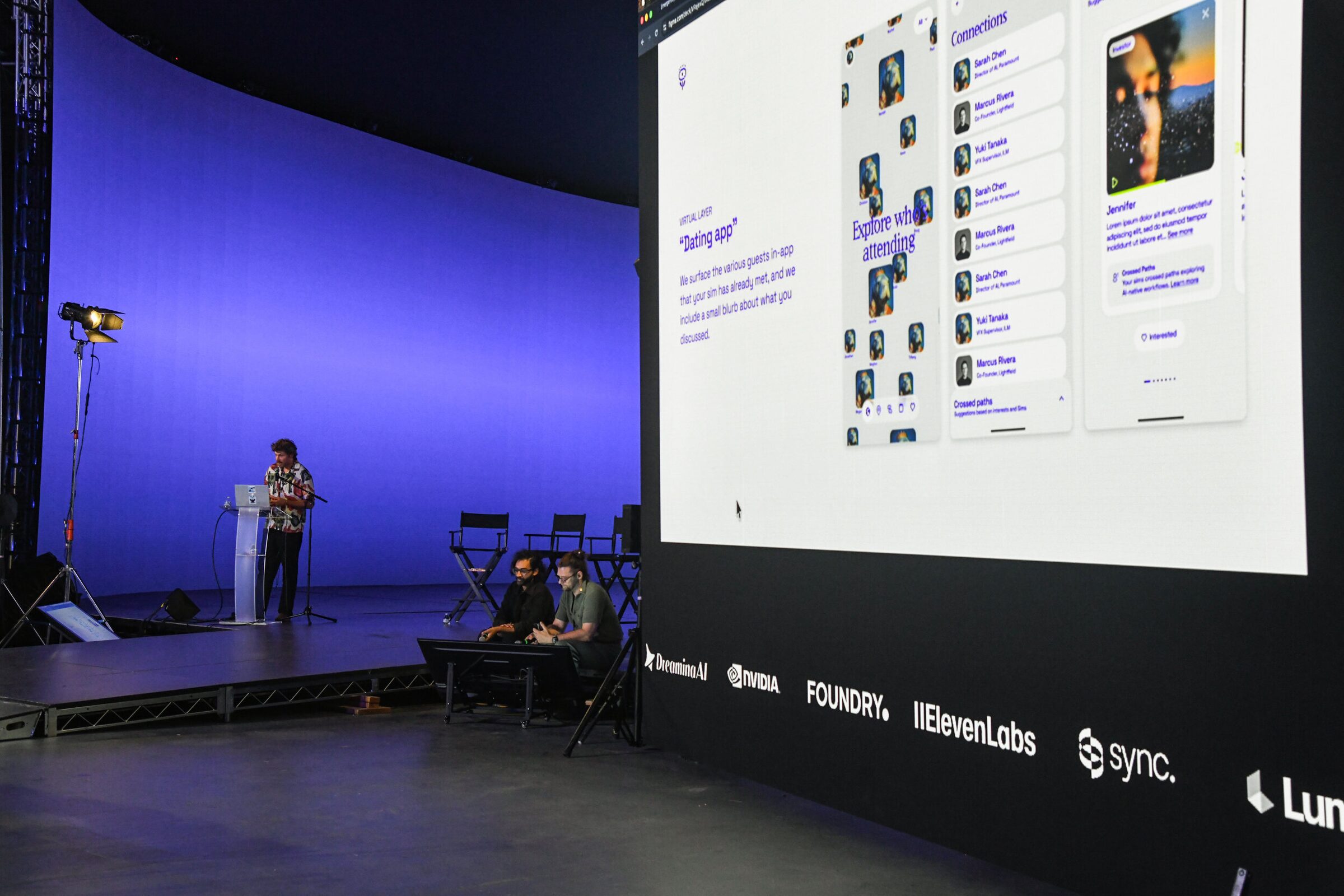 Ian Eck speaks at a lectern on a blue curved stage while a giant screen beside him shows a simulated attendee app.